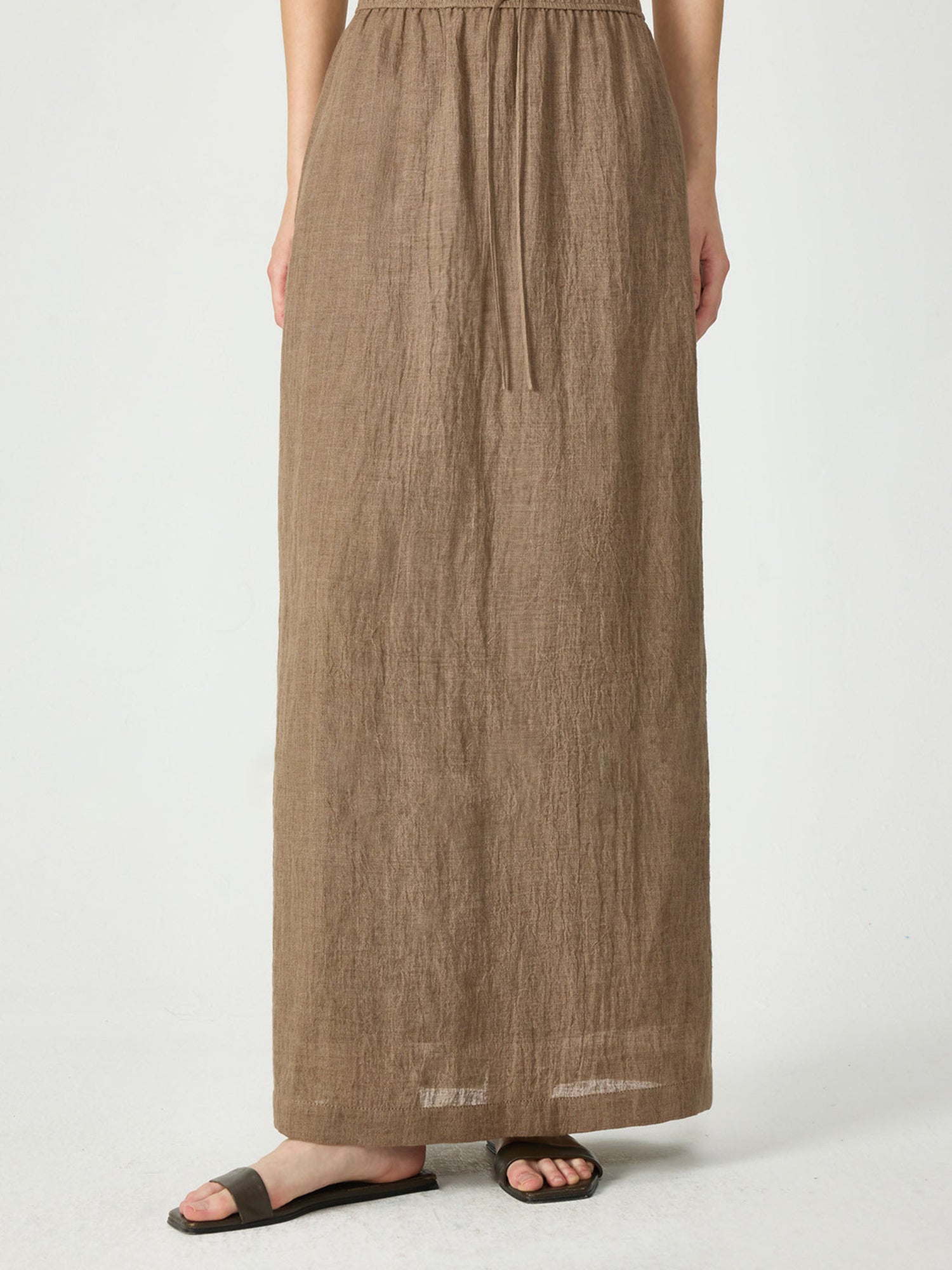 Woman wearing Arkin Studio crinkled linen relaxed midi skirt in coffee, with a pair of brown sandals, standing facing the camera, on a white background