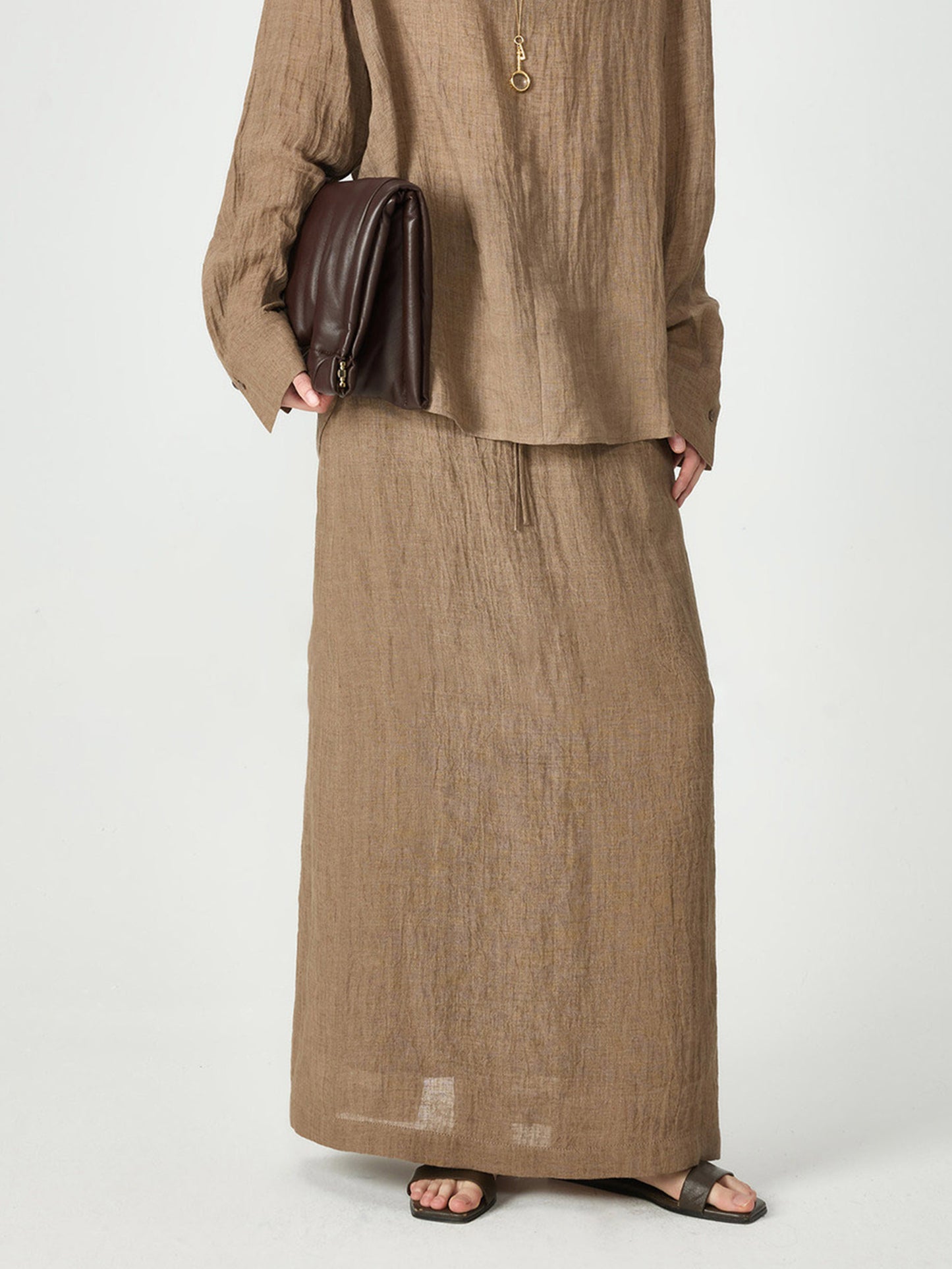 Woman wearing Arkin Studio crinkled linen relaxed midi skirt in coffee, holding a brown bag, with a pair of brown sandals, standing facing the camera, on a white background