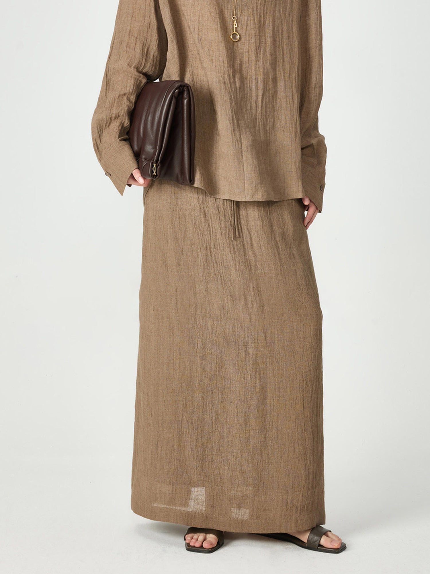 Woman wearing Arkin Studio crinkled linen relaxed midi skirt in coffee, holding a brown bag, with a pair of brown sandals, standing facing the camera, on a white background
