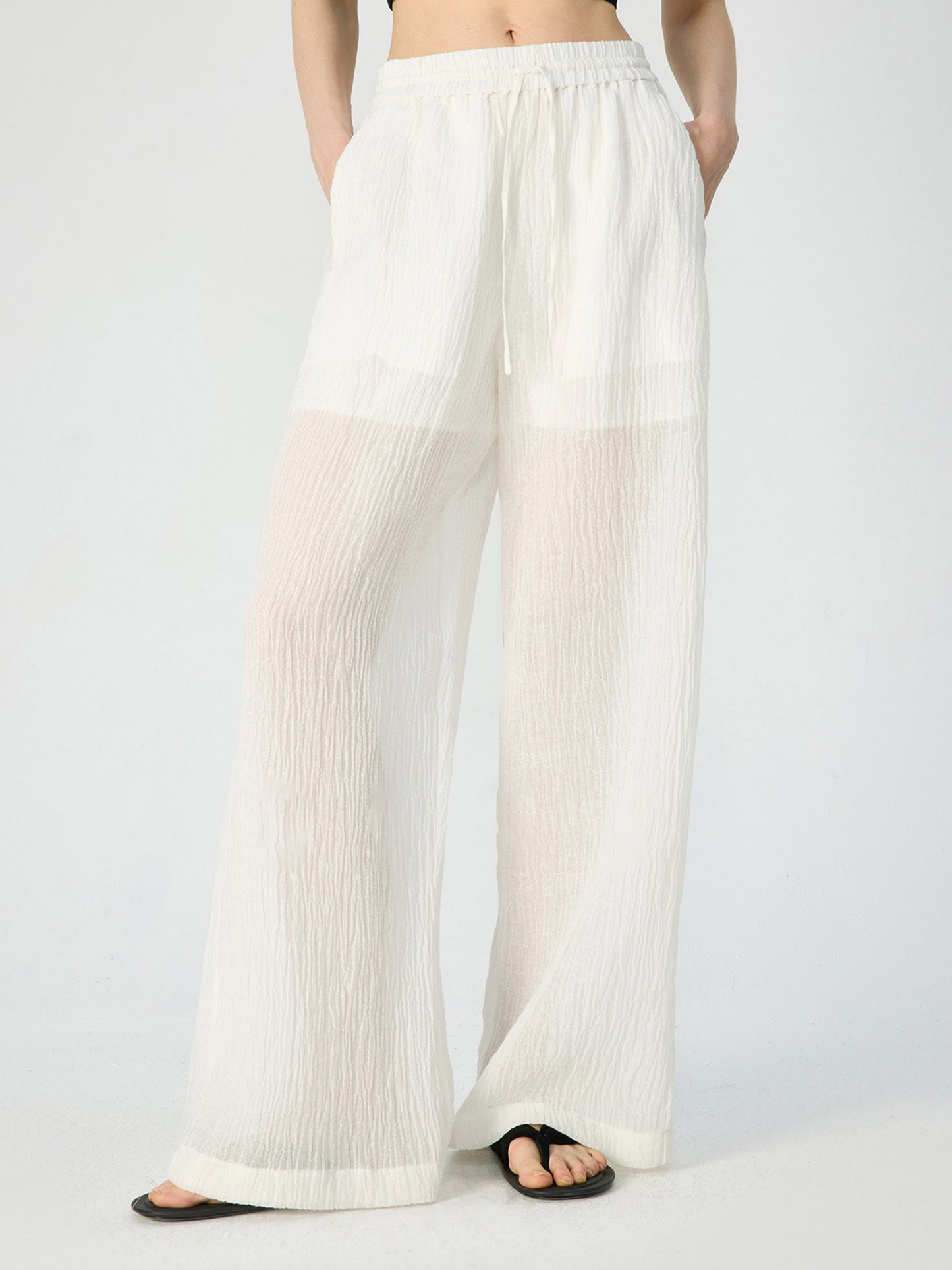 Woman wearing Arkin Studio crinkled linen wide leg pants in ivory, with a pair of black flip-flops, hands in the pants’ pockets, standing facing the camera, on a white background