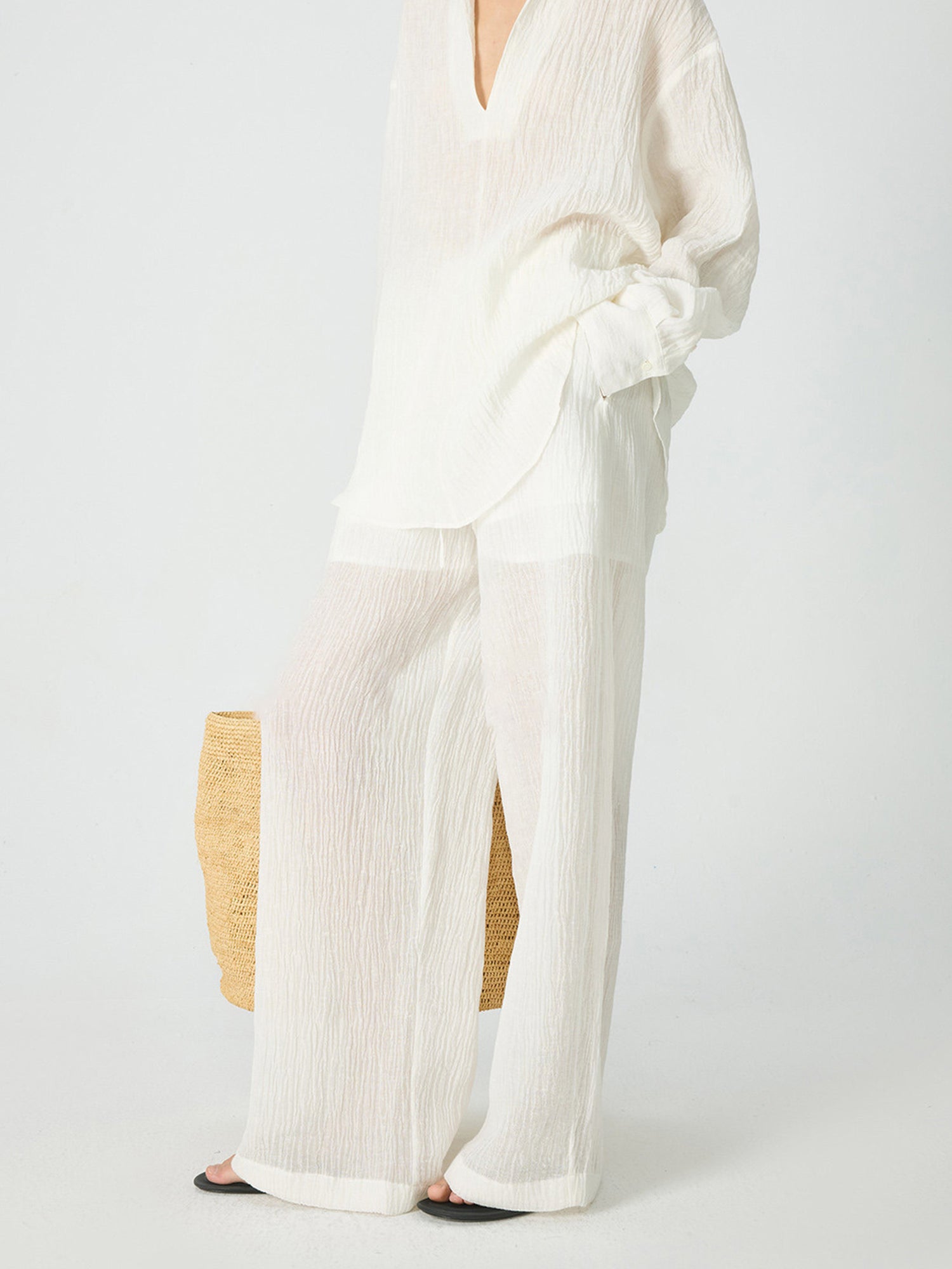 Woman wearing Arkin Studio crinkled linen wide leg pants in ivory, with a pair of brown flip-flops, one hand in a pants’ pocket, another hand hold a big woven bag, standing sideways, on a white background