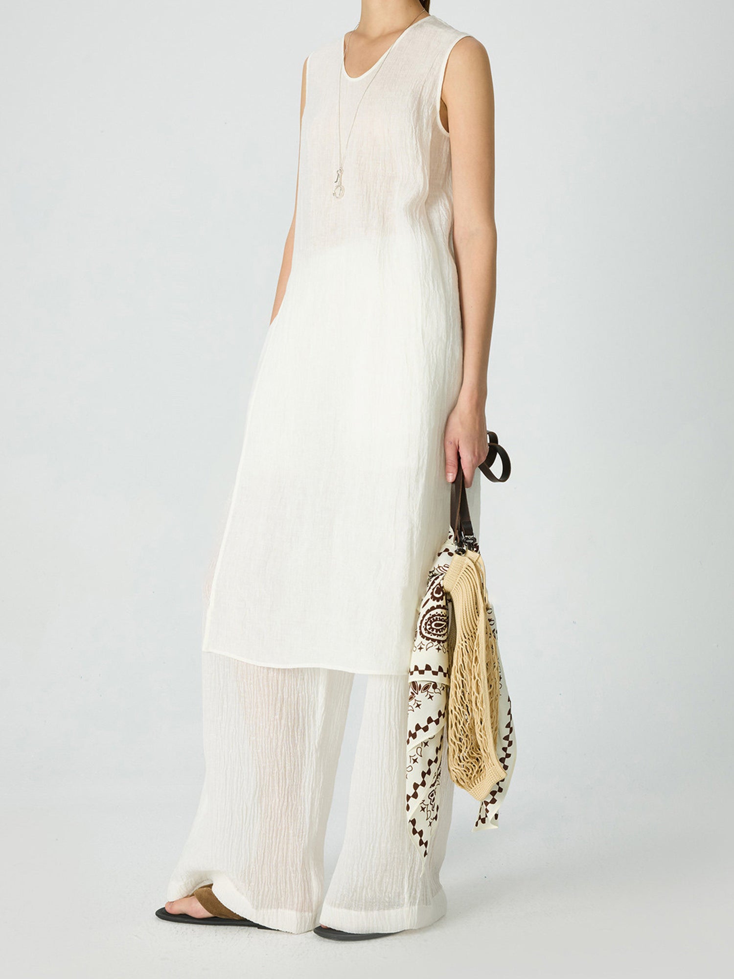 Woman wearing Arkin Studio crinkled linen wide leg pants in ivory, with a pair of brown flip-flops, holding a bag in one hand, standing sideways, on a white background