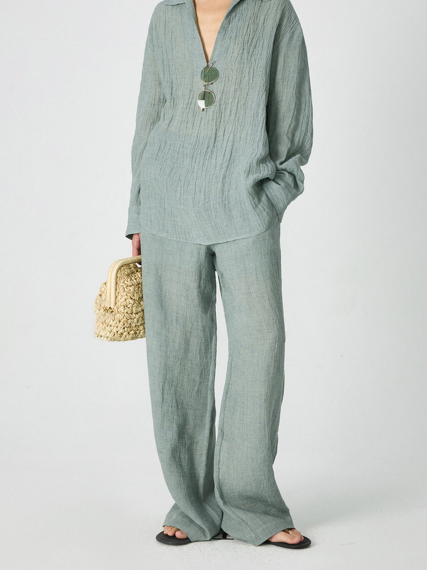 Woman wearing Arkin Studio crinkled linen wide leg pants in mint-green, with a pair of glasses on the shirt, with a pair of brown sandals, holding a small woven bag, standing facing the camera, on a white background