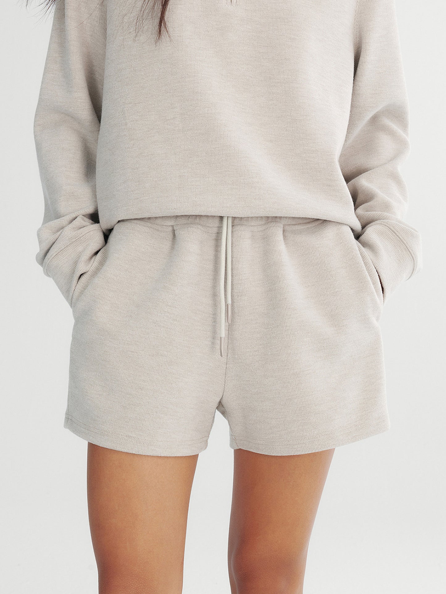 Woman wearing Arkin Studio drawstring high waist sport shorts in light gray and matching top, hands in the pockets, standing facing the camera, on a white background