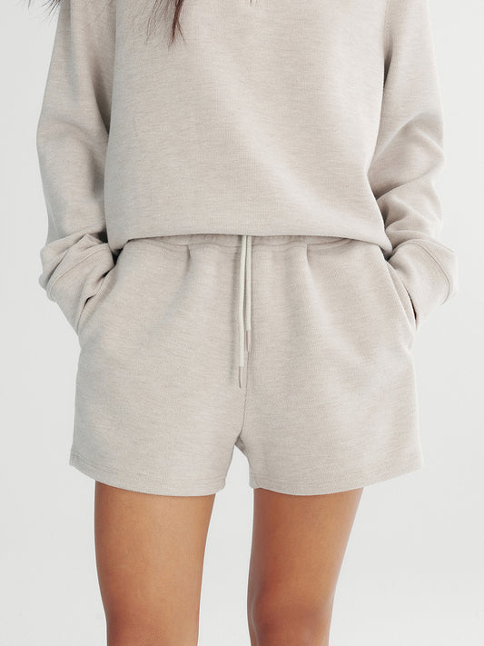 Woman wearing Arkin Studio drawstring high waist sport shorts in light gray and matching top, hands in the pockets, standing facing the camera, on a white background
