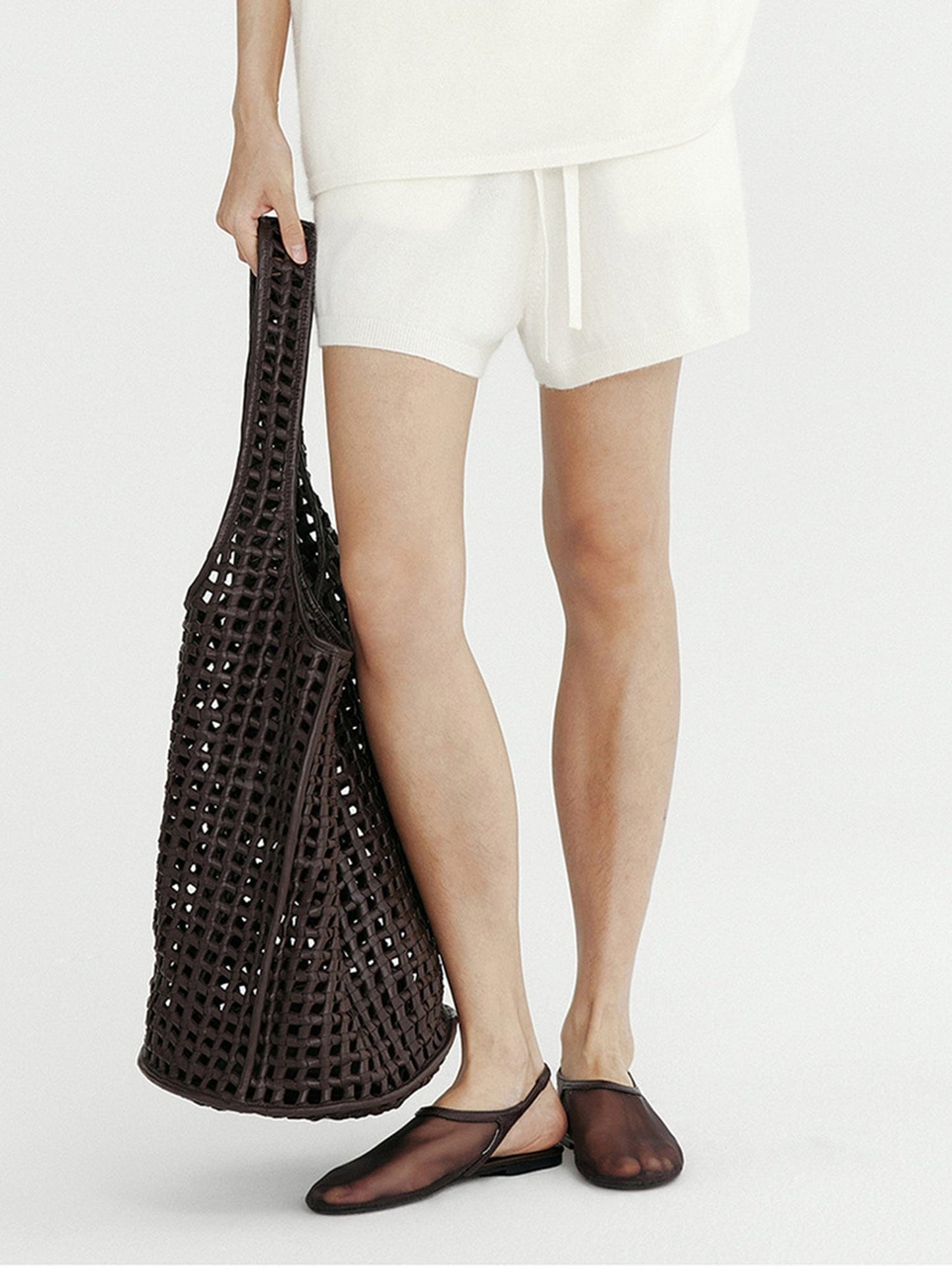 Woman wearing Arkin Studio ribbed knit drawstring shorts in white and matching top, holding a chocolate bag, wearing a pair of chocolate shoes, standing facing the camera, on a white background