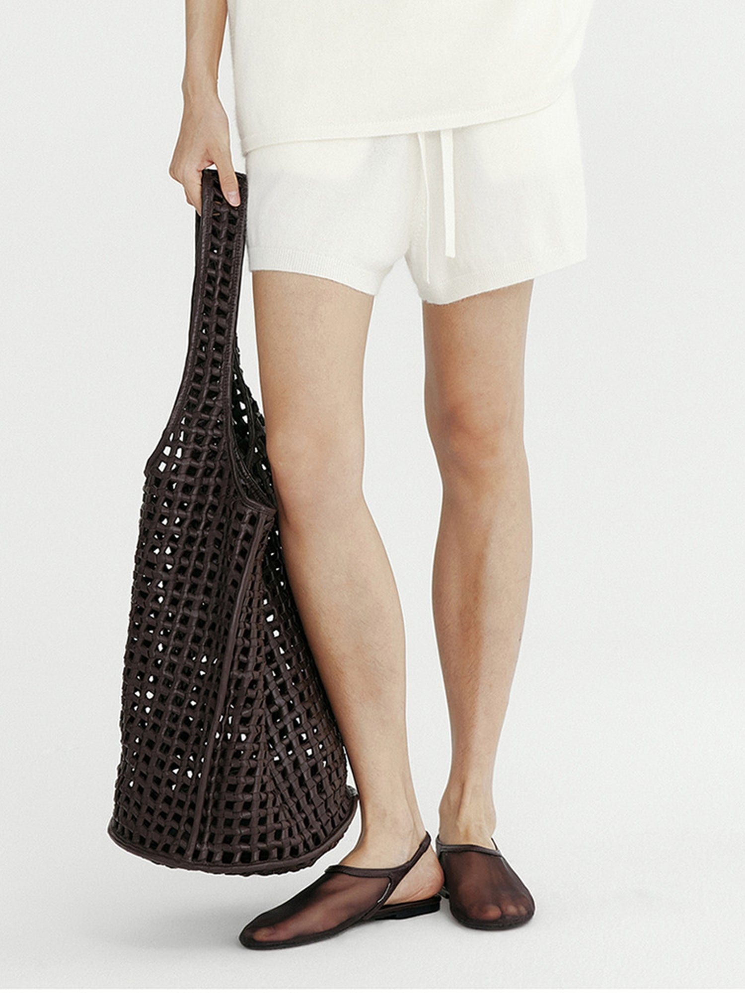 Woman wearing Arkin Studio ribbed knit drawstring shorts in white and matching top, holding a chocolate bag, wearing a pair of chocolate shoes, standing facing the camera, on a white background