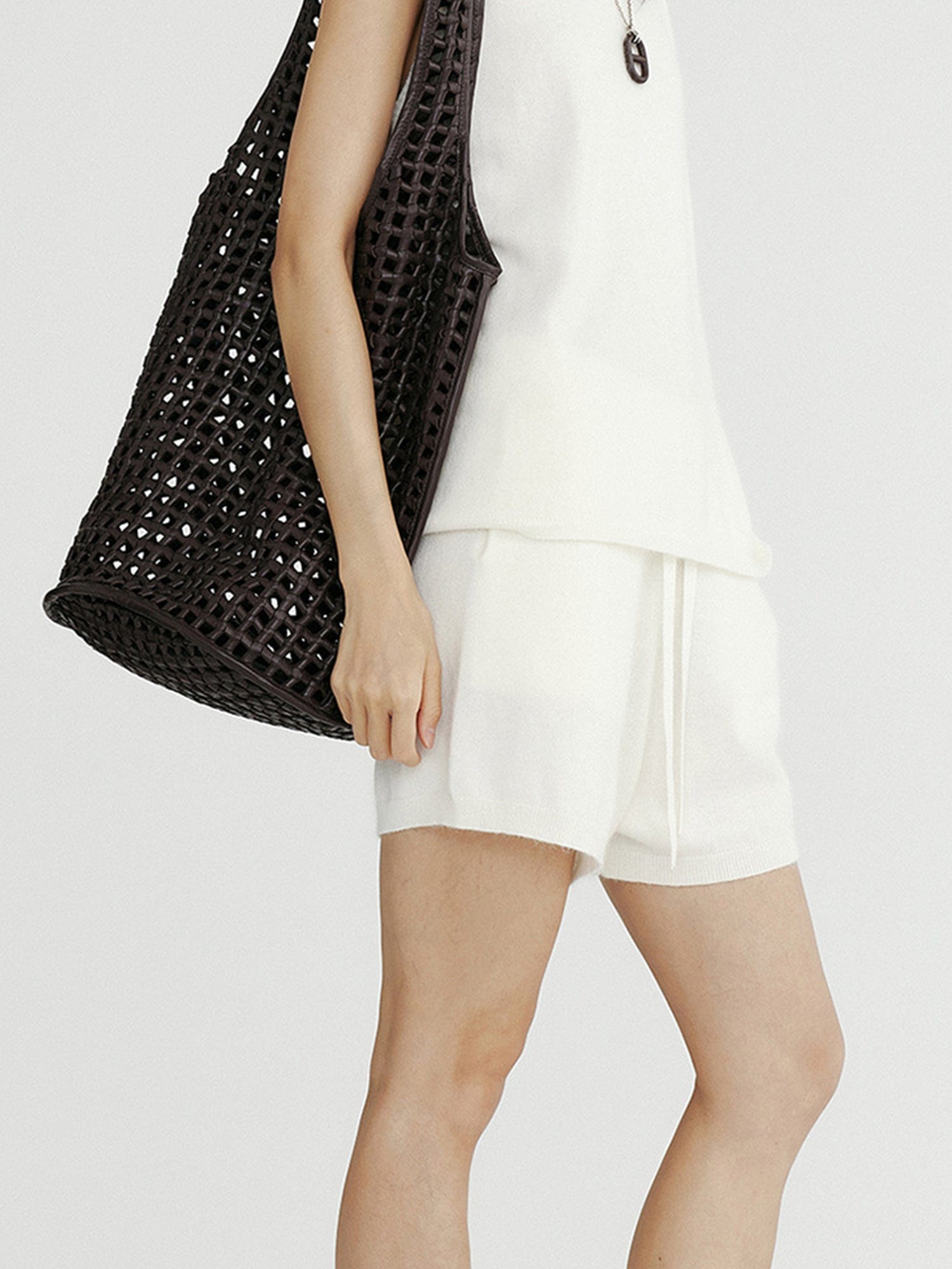 Woman wearing Arkin Studio ribbed knit drawstring shorts in white and matching top, a chocolate bag on her right shoulder, standing sideways, on a white background