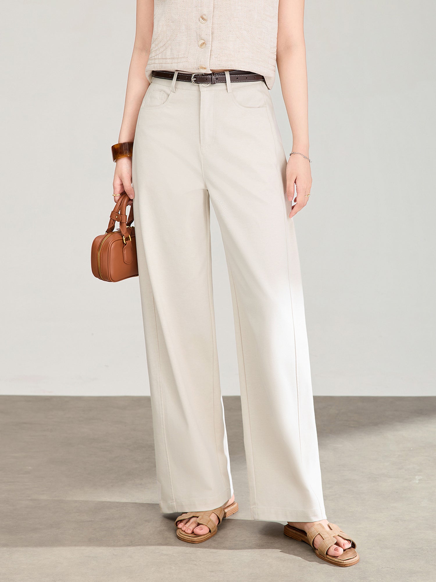 Woman wearing Arkin Studio high rise curved leg trousers in ivory with a brown belt, and a beige vest, holding a brown handbag, standing in front of a wall, facing the camera, on a gray floor