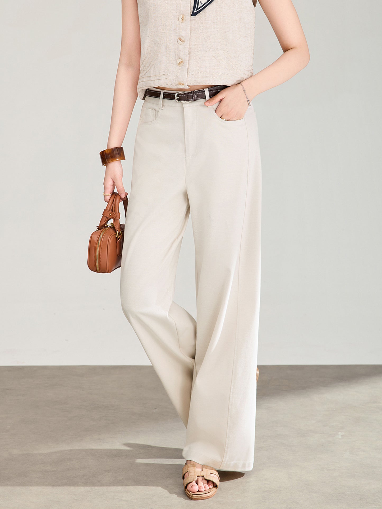 Woman wearing Arkin Studio high rise curved leg trousers in ivory with a brown belt, and a beige vest, holding a brown handbag, standing in front of a wall, facing the camera, with her right toe tapping the ground, on a gray floor