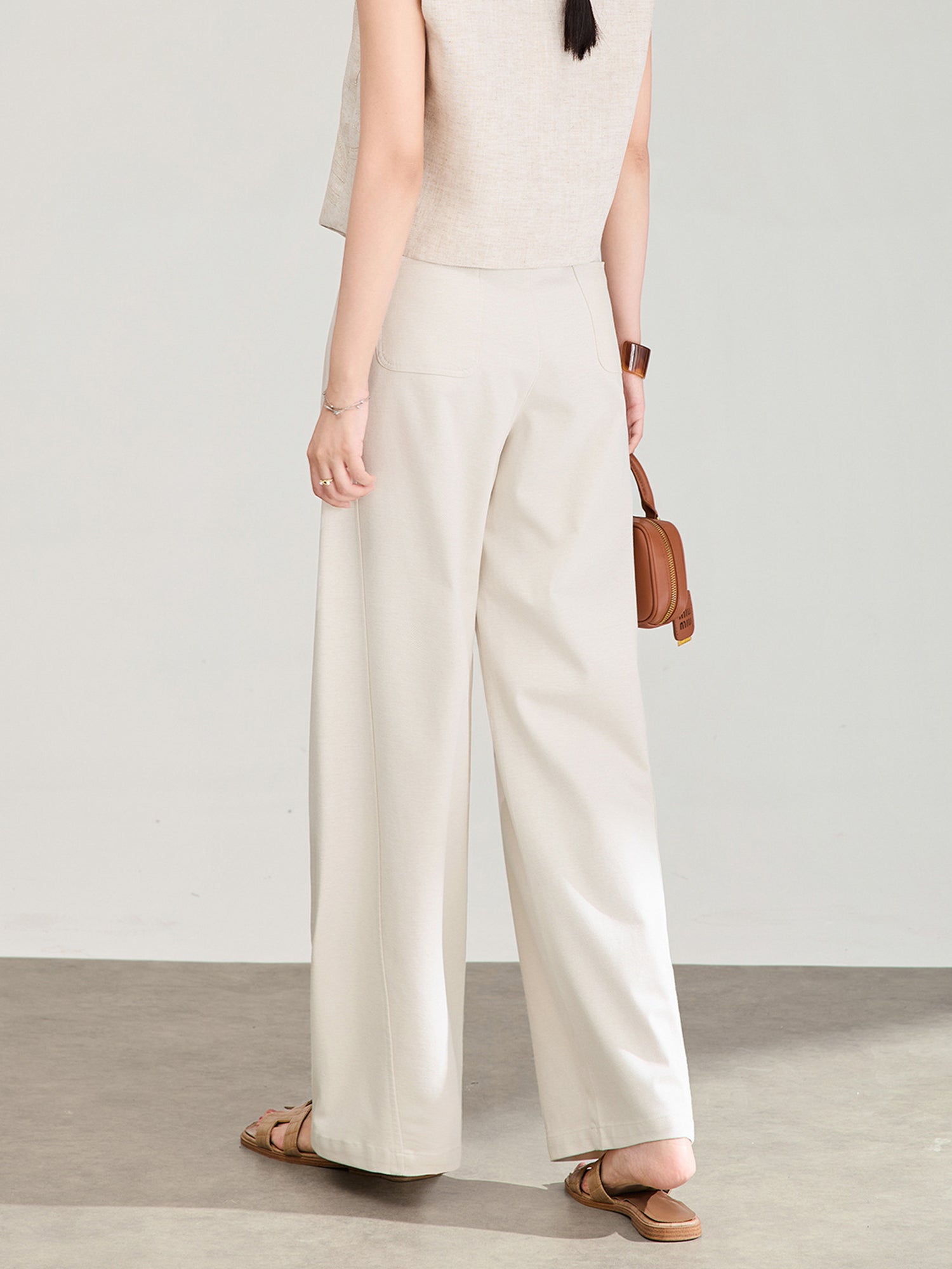 Woman wearing Arkin Studio high rise curved leg trousers in ivory with a brown belt, and a beige vest, with a pair of slippers, holding a brown handbag, standing facing the wall, on a gray floor