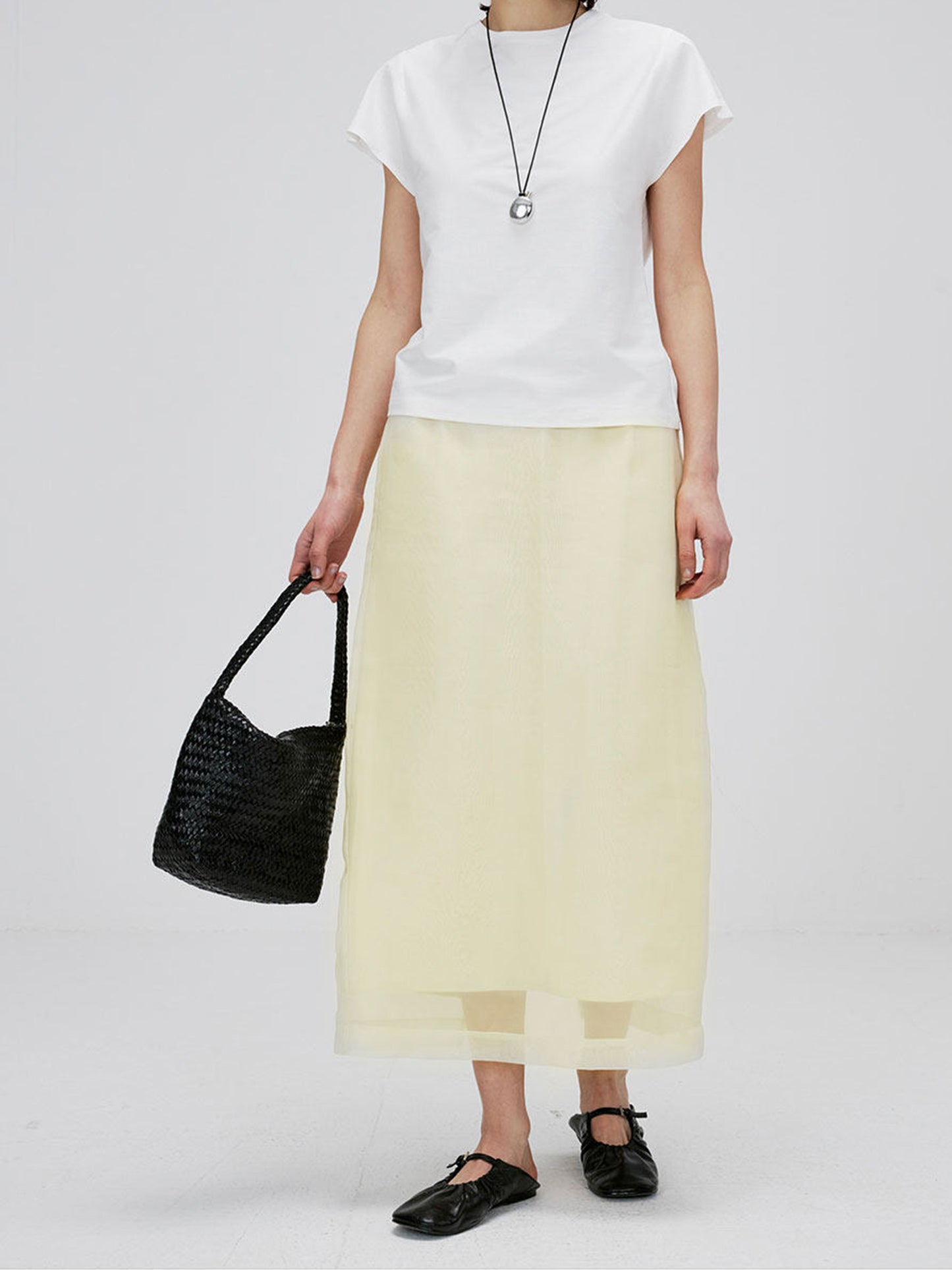 Woman wearing Arkin Studio high-waist mesh two-layer midi skirt in light yellow and a white short sleeve, with a black bag in a hand, standing facing camera, on white background
