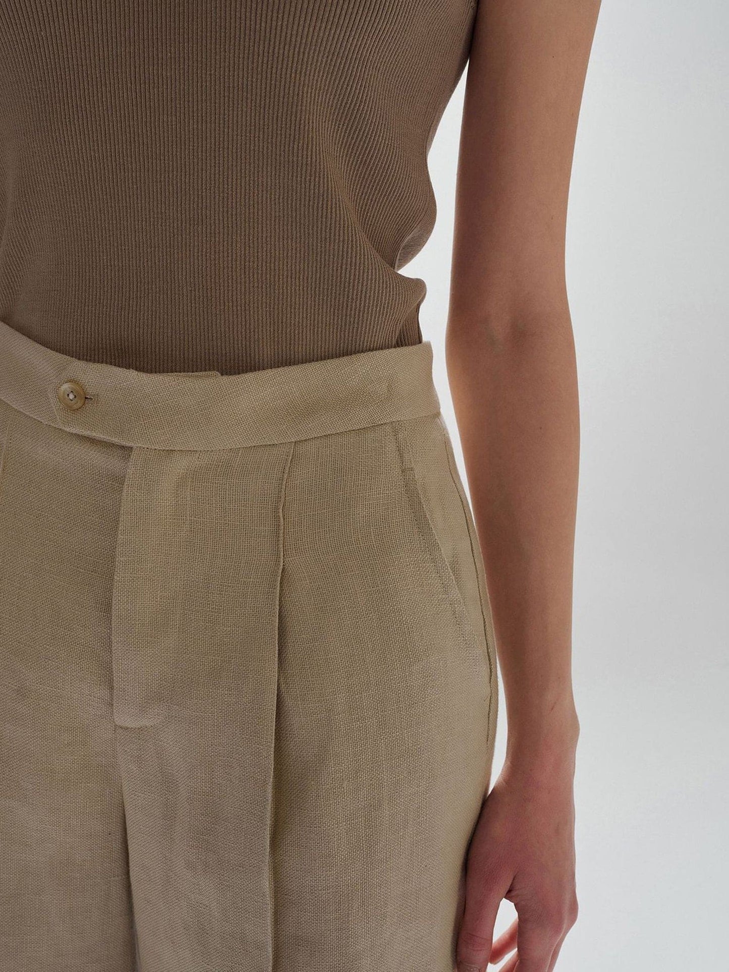 Close up of a woman's waist wearing Arkin Studio high waist turn up linen shorts in khaki and a brown top, standing with arms down at her sides, facing the camera, on a white background