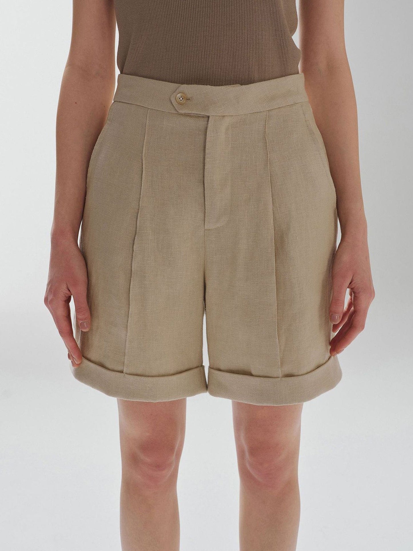 Woman wearing Arkin Studio high waist turn up linen shorts in khaki and a brown top, standing with arms down at her sides, facing the camera, on a white background