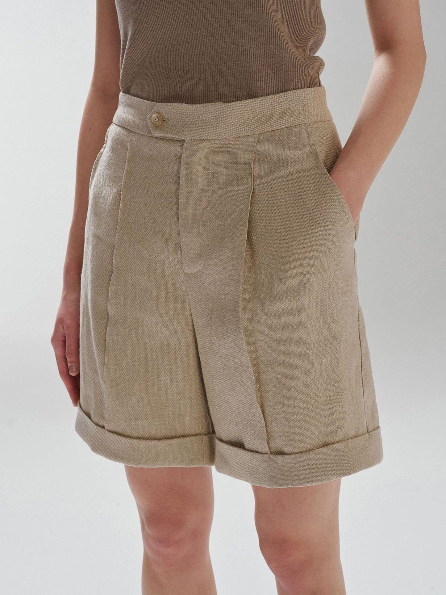 Woman wearing Arkin Studio high waist turn up linen shorts in khaki and a brown top, standing sideways with one hand in a pocket, on a white background
