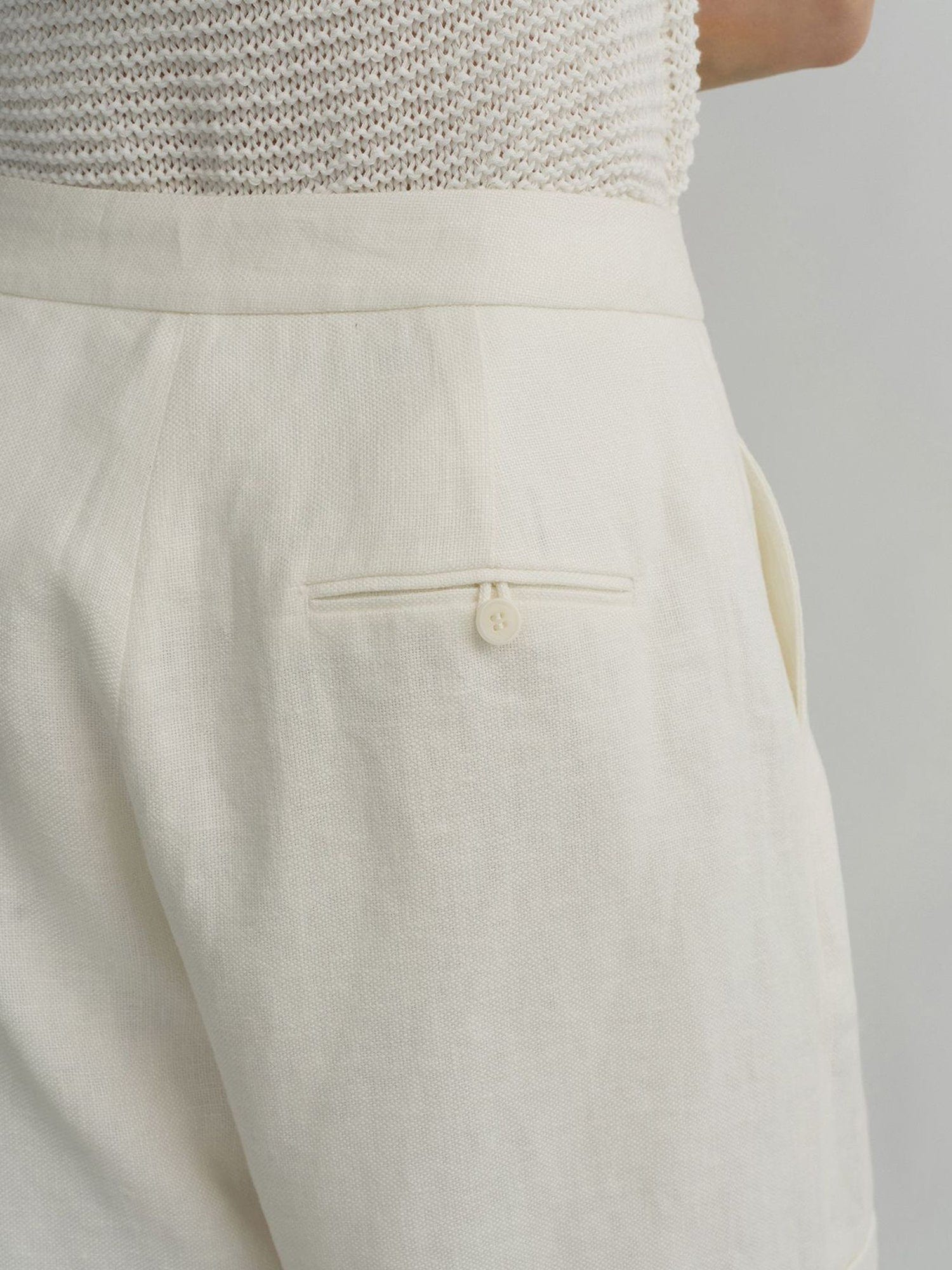 Woman wearing Arkin Studio high waist turn up linen shorts in white and a white top, standing facing a white wall, close up of her back and waist, on a white background