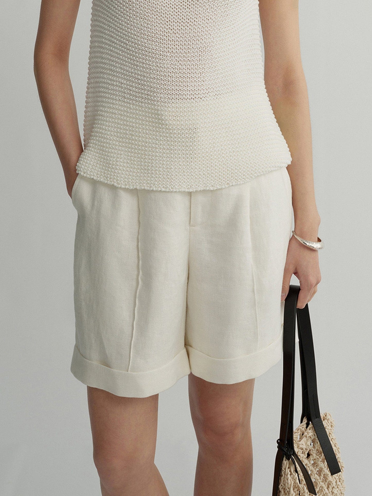 Woman wearing Arkin Studio high waist turn up linen shorts in white and a white top, standing with a woven bag in left hand, facing the camera, on a white background
