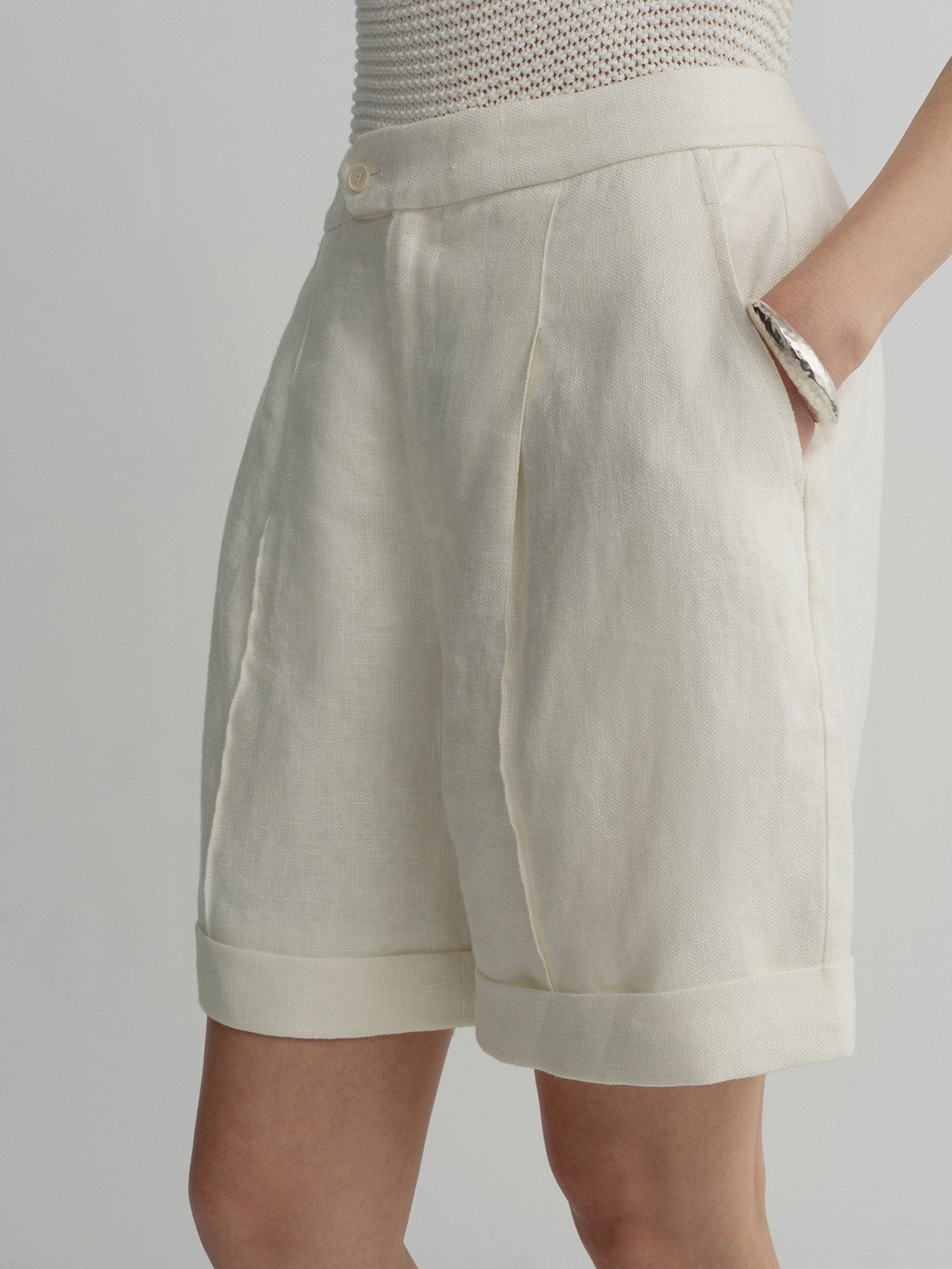 Woman wearing Arkin Studio high waist turn up linen shorts in white and a white top, standing sideways with one hand in a pocket, on a white background