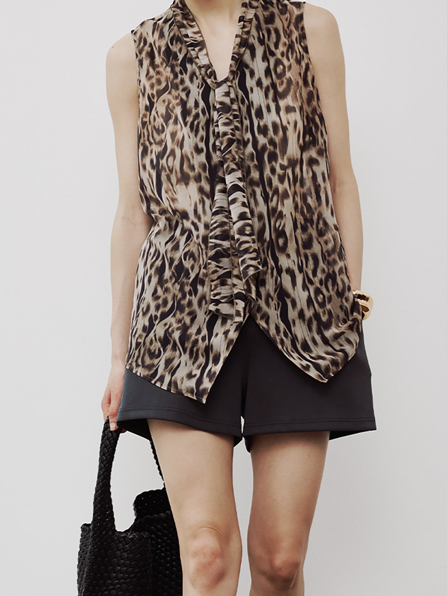 Woman wearing Arkin Studio leopard chiffon v-neck tie blouse in brown and black shorts, with a black handbag, standing facing the camera, on a white background