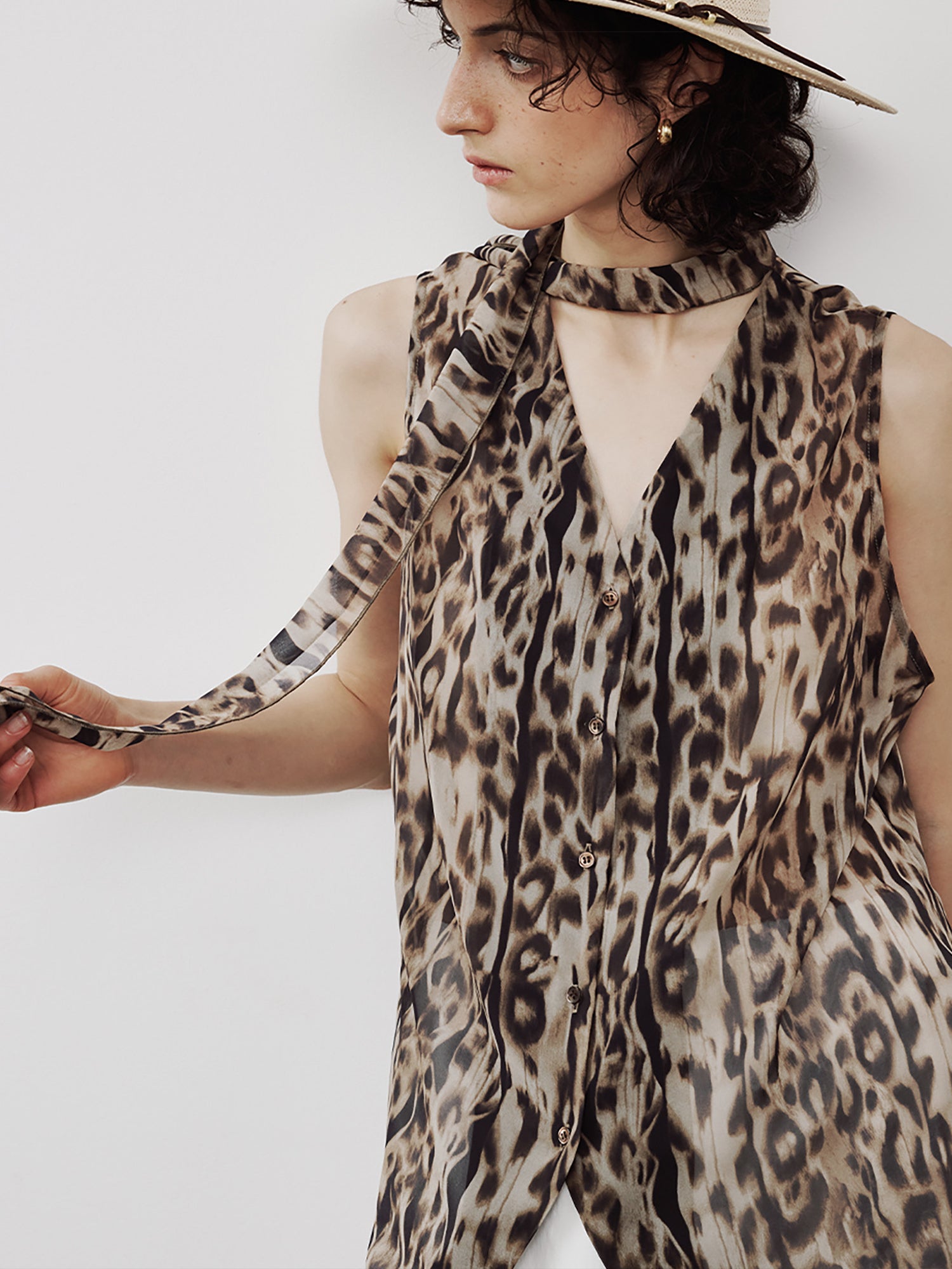 Woman wearing Arkin Studio leopard chiffon v-neck tie blouse in brown and white pants, head turned to the right, holding a long strap of the blouse in her hand, on a white background