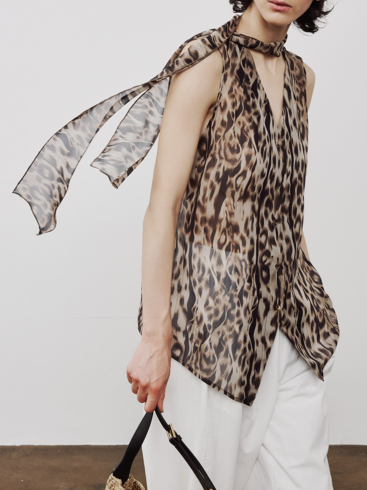 Woman wearing Arkin Studio leopard chiffon v-neck tie blouse in brown and white pants, head turned to the right, holding a handbag, standing sideways, on a white background