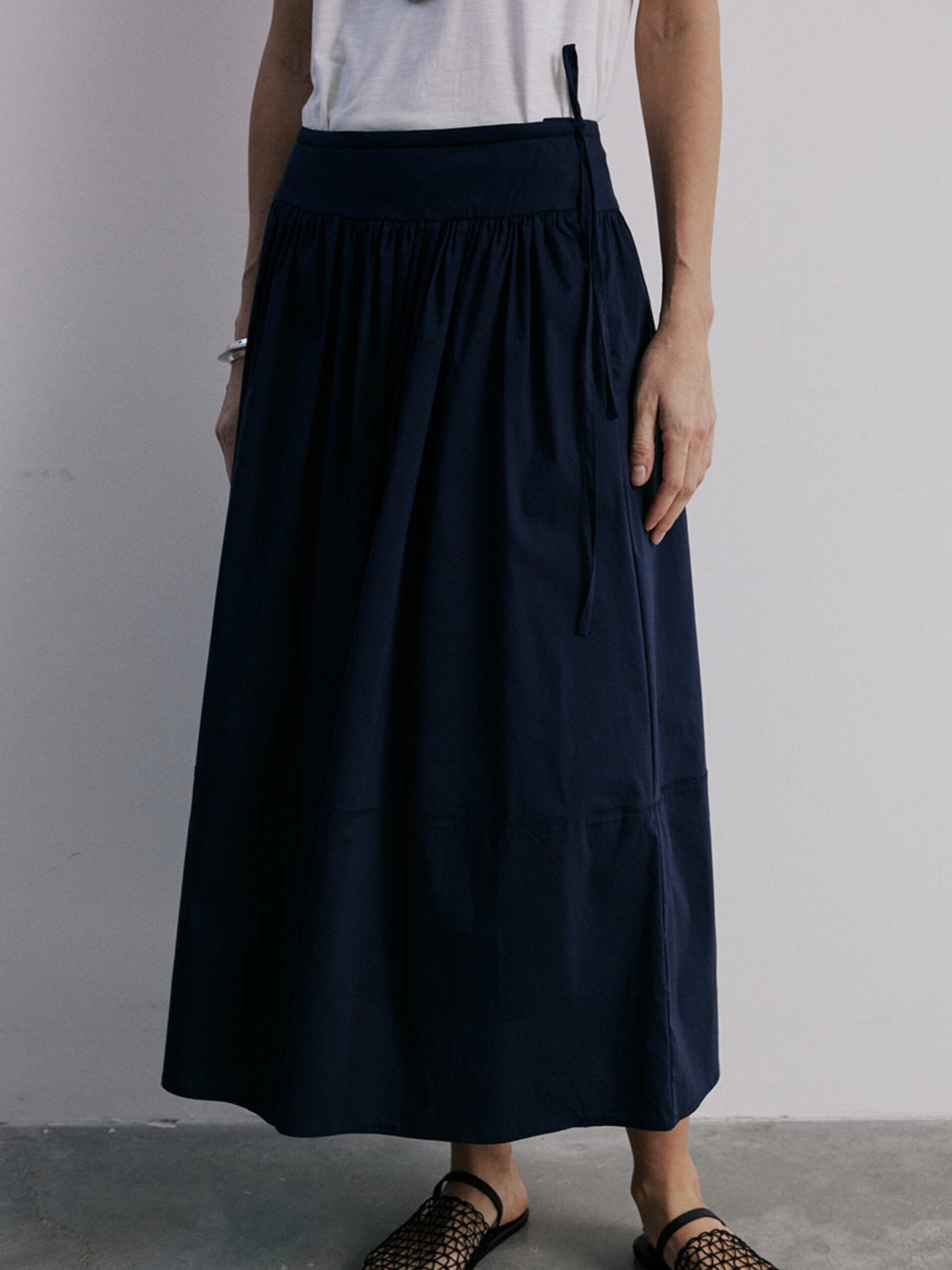 Woman wearing Arkin Studio lightweight pleated a-line drawstring skirt in dark-navy and a white sleeveless top, facing the camera on the cement floor, a white wall behind her