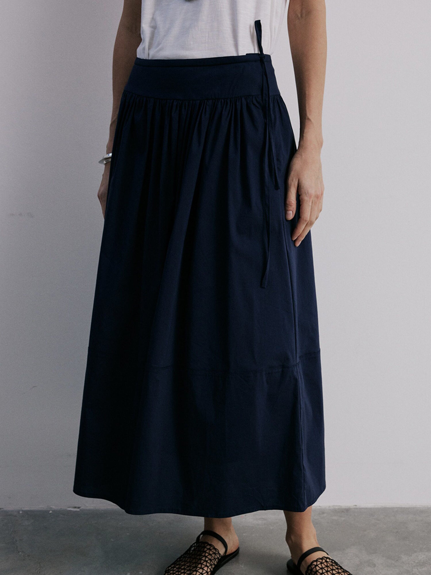 Woman wearing Arkin Studio lightweight pleated a-line drawstring skirt in dark-navy and a white sleeveless top, facing the camera on the cement floor, a white wall behind her