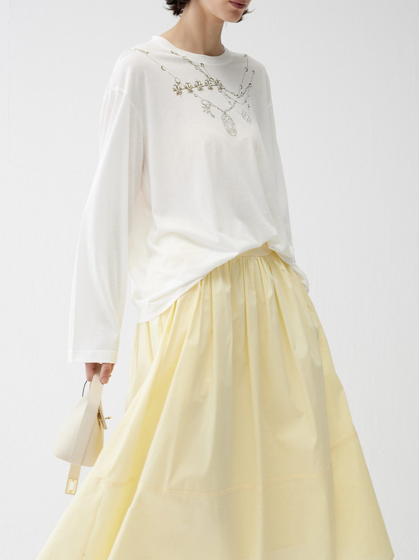 Woman wearing Arkin Studio lightweight pleated a-line drawstring skirt in light-yellow and a white blouse with floral embroidery, with a white bag in hand, three-quarter view, on a white background
