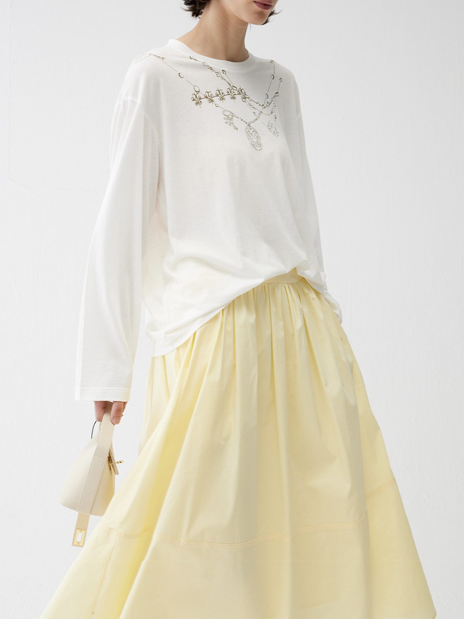 Woman wearing Arkin Studio lightweight pleated a-line drawstring skirt in light-yellow and a white blouse with floral embroidery, with a white bag in hand, three-quarter view, on a white background