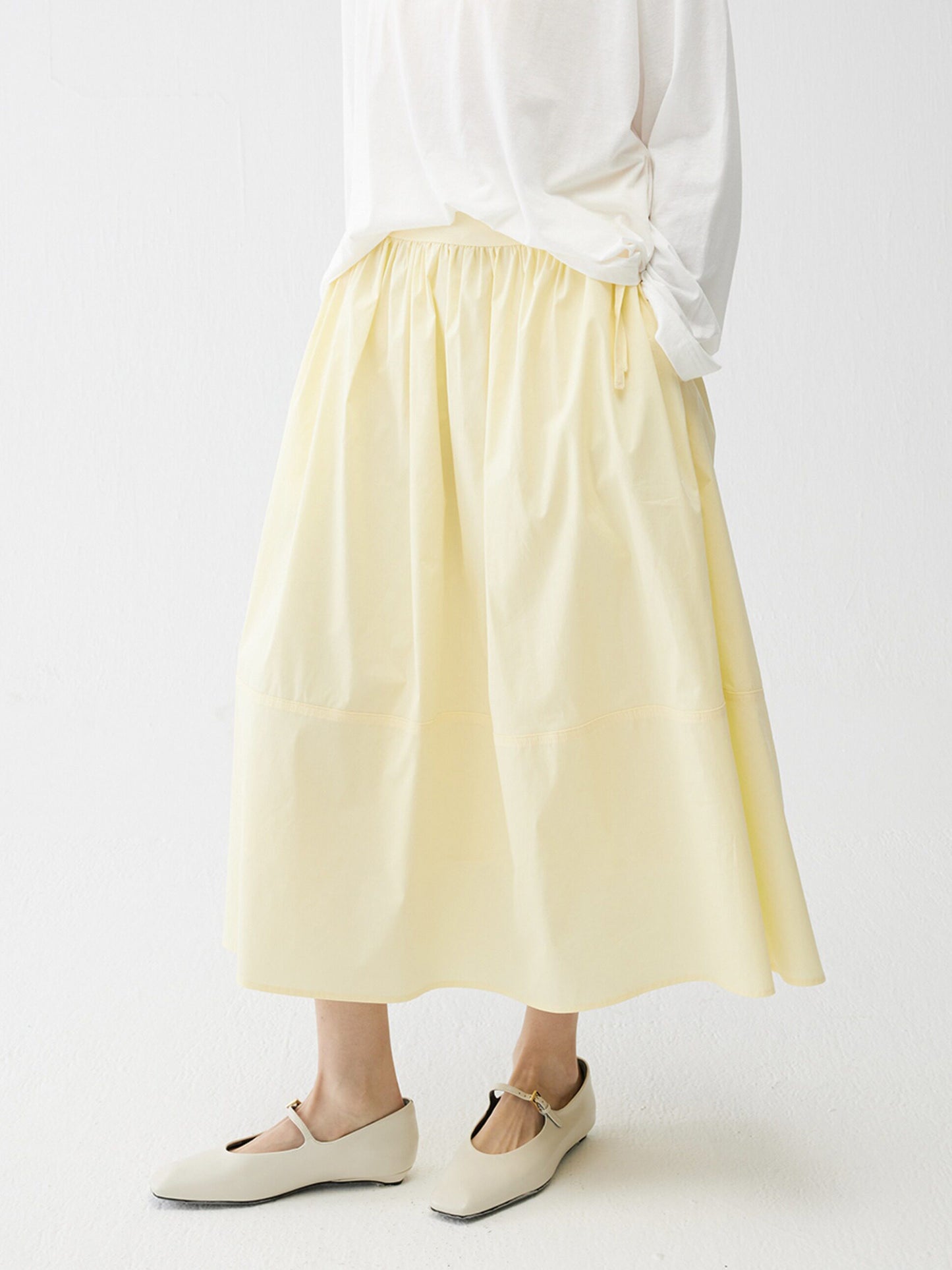 Woman wearing Arkin Studio lightweight pleated a-line drawstring skirt in light-yellow and a white blouse, with summer sandals on feet, three-quarter view, on a white background