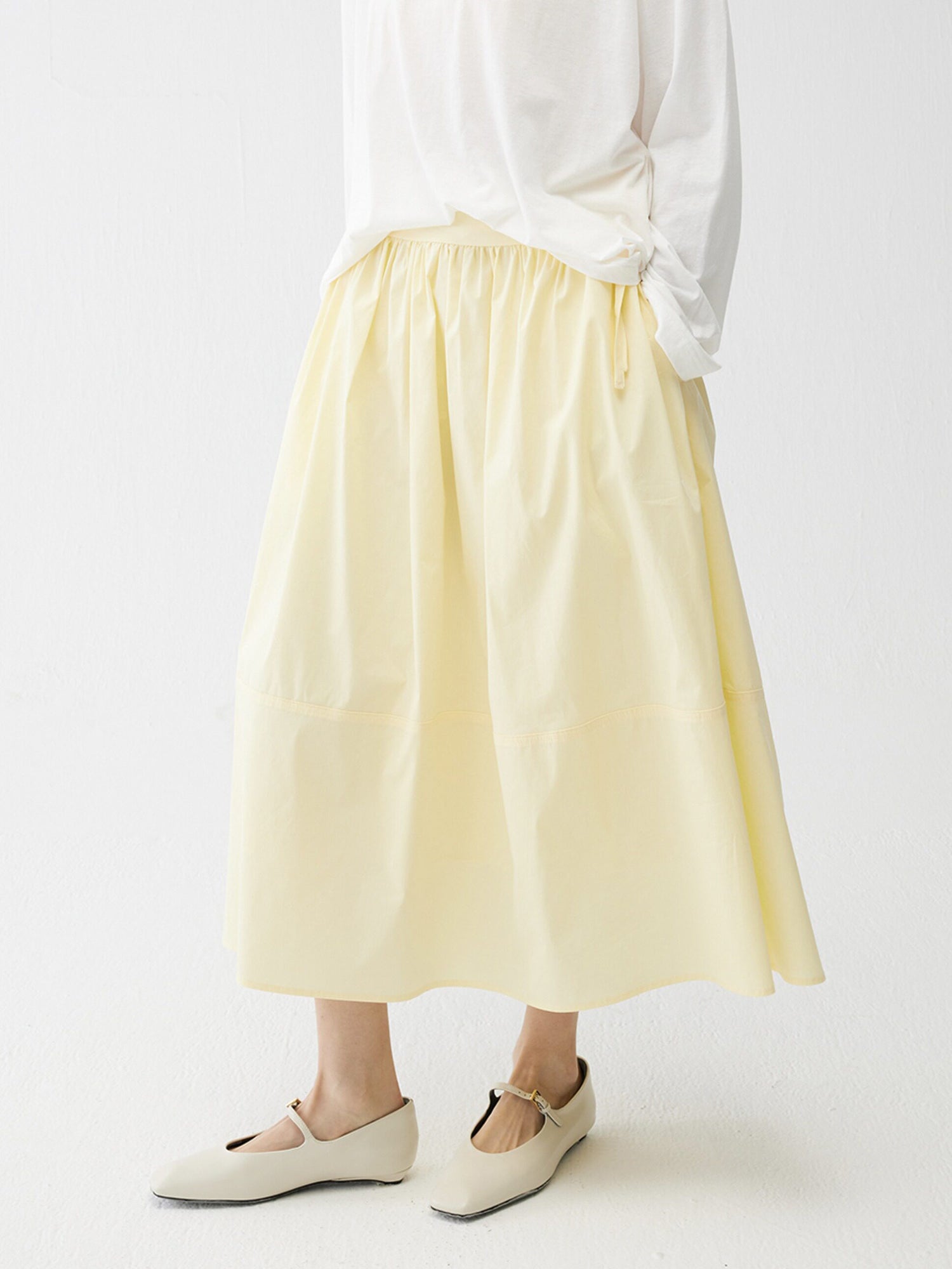 Woman wearing Arkin Studio lightweight pleated a-line drawstring skirt in light-yellow and a white blouse, with summer sandals on feet, three-quarter view, on a white background