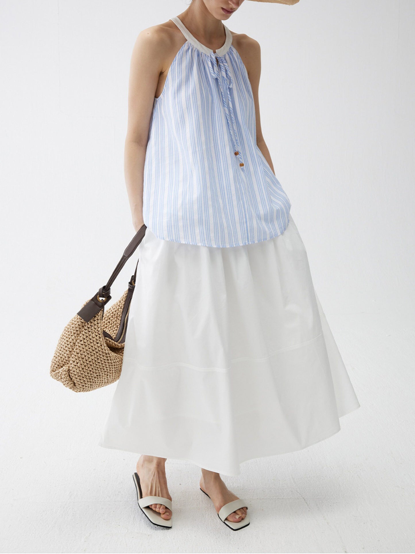 Woman wearing a blue striped top and Arkin Studio lightweight pleated a-line drawstring skirt in white, with a woven bag , head down and three-quarter view, on a white background