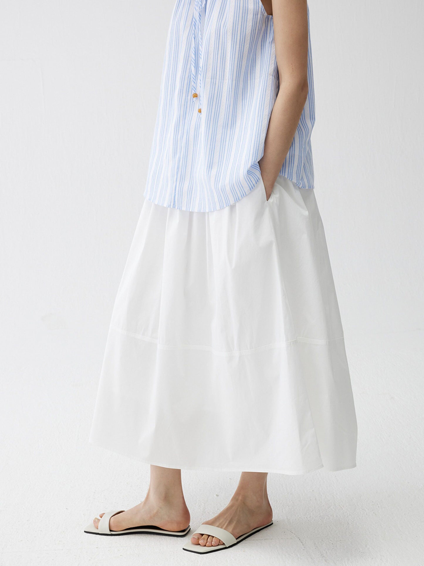 Woman wearing a blue striped top and Arkin Studio lightweight pleated a-line drawstring skirt in white, standing full side view with hand in pocket, on a white background