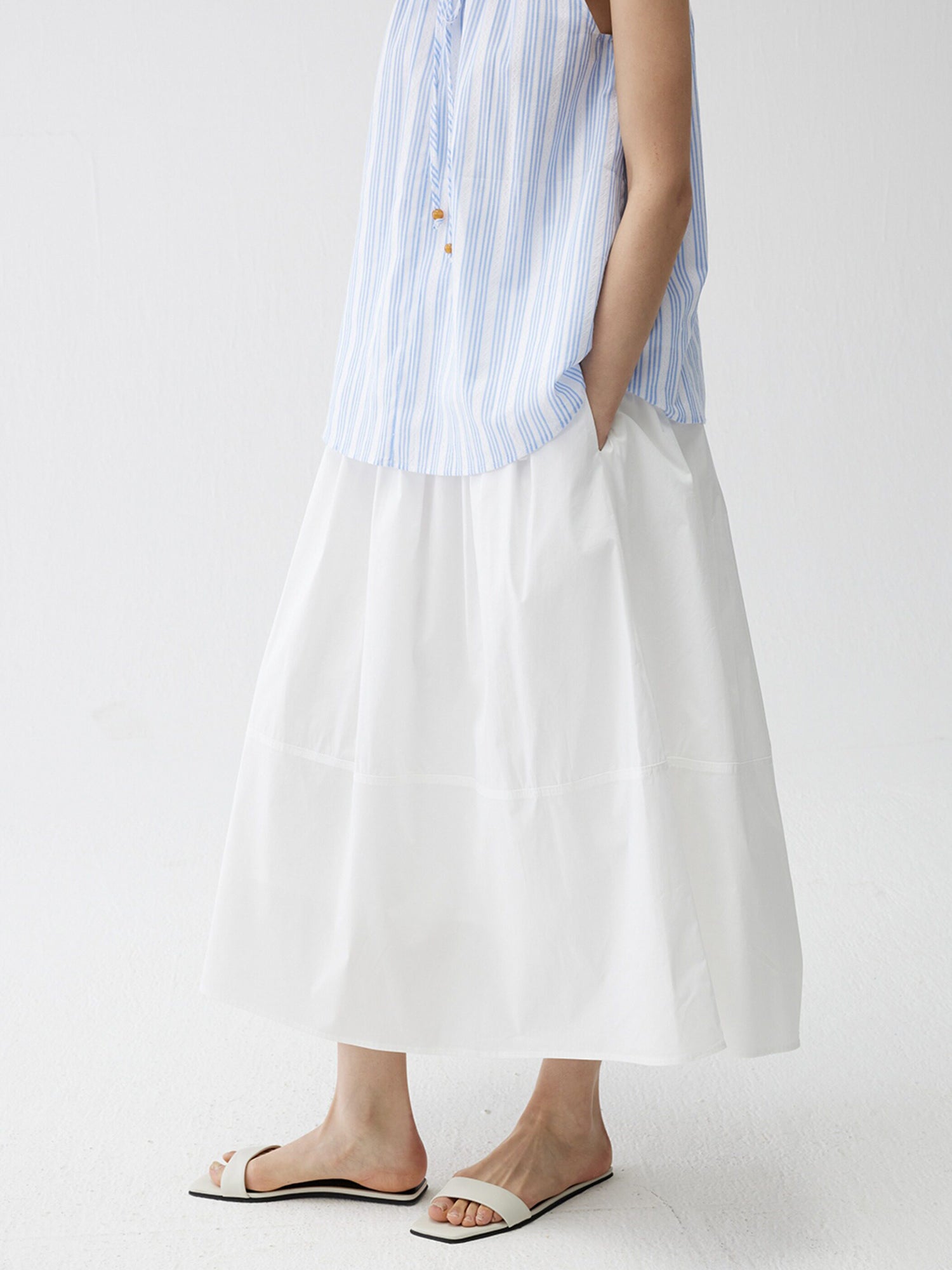 Woman wearing a blue striped top and Arkin Studio lightweight pleated a-line drawstring skirt in white, standing full side view with hand in pocket, on a white background