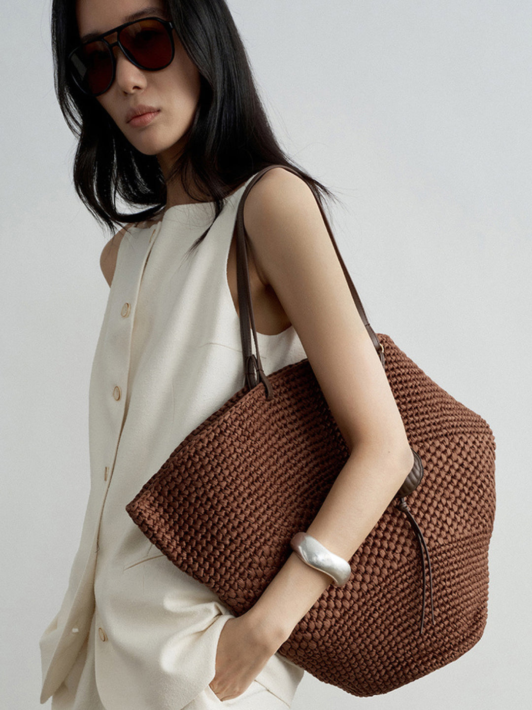 Lookbook: Woman wearing Arkin Studio boatneck slim strap button front vest in ivory and matching shorts, a woven brown bag on her left shoulder, a silver bracelet on her left hand, wearing sunglasses, standing sideways in front of a white background – shop the lookbook