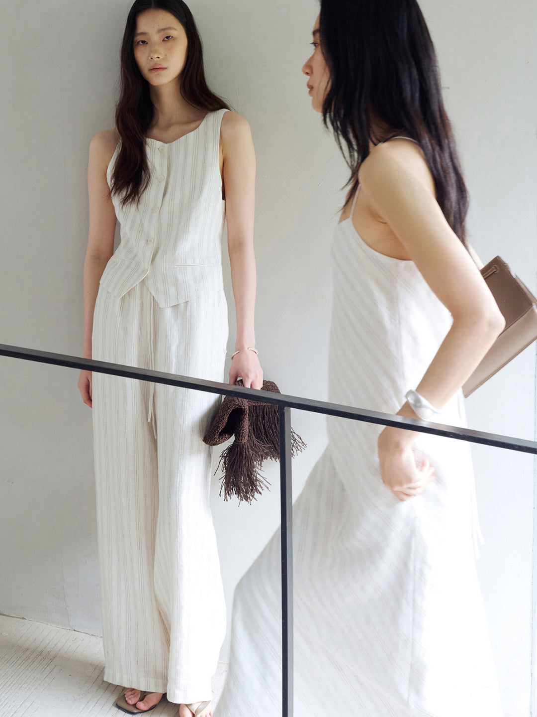 Lookbook: Left model wearing Arkin Studio ivory striped linen drawstring wide leg pants and matching striped sleeveless vest, holding a brown handbag, standing facing the camera. Right model wearing a white striped dress, standing sideways - shop the lookbook