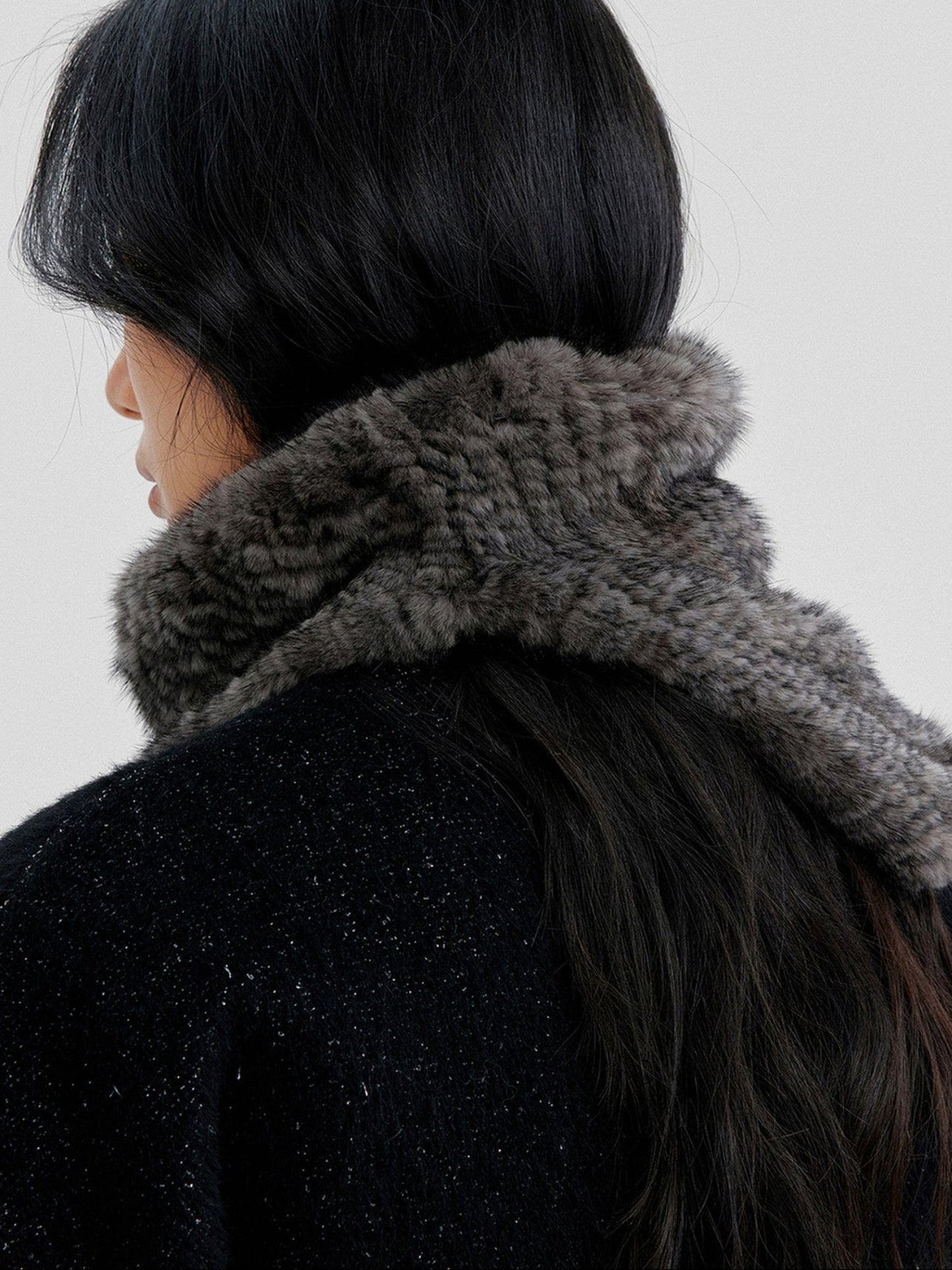Close-up of a woman's upper back and back of head wearing Arkin Studio metallic trim belted wool coat in black with a furry scarf, with long hair, standing facing a white wall, on a white background