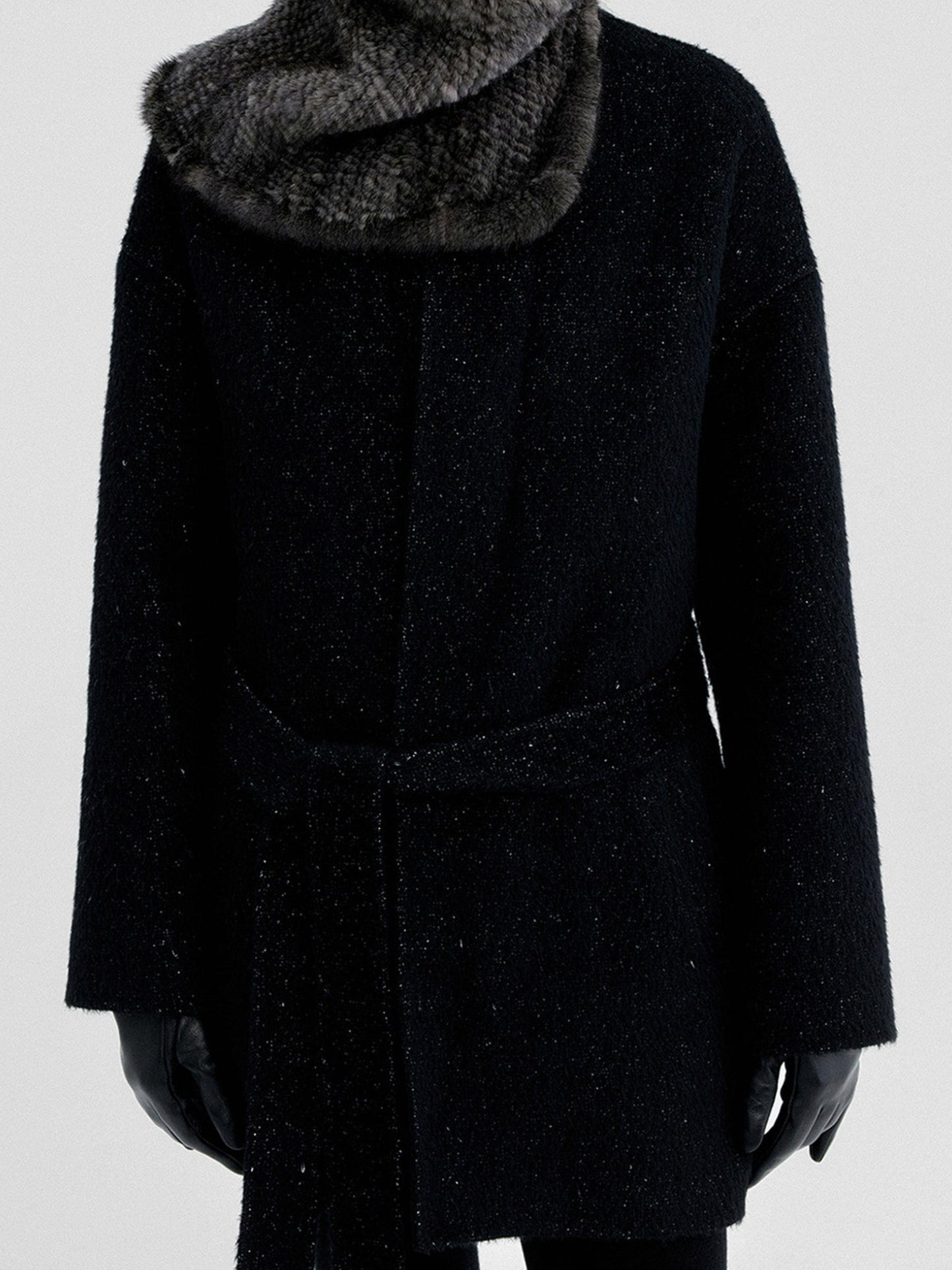 Woman wearing Arkin Studio metallic trim belted wool coat in black with furry scarf, hands down at her sides, standing facing the camera, on a white background