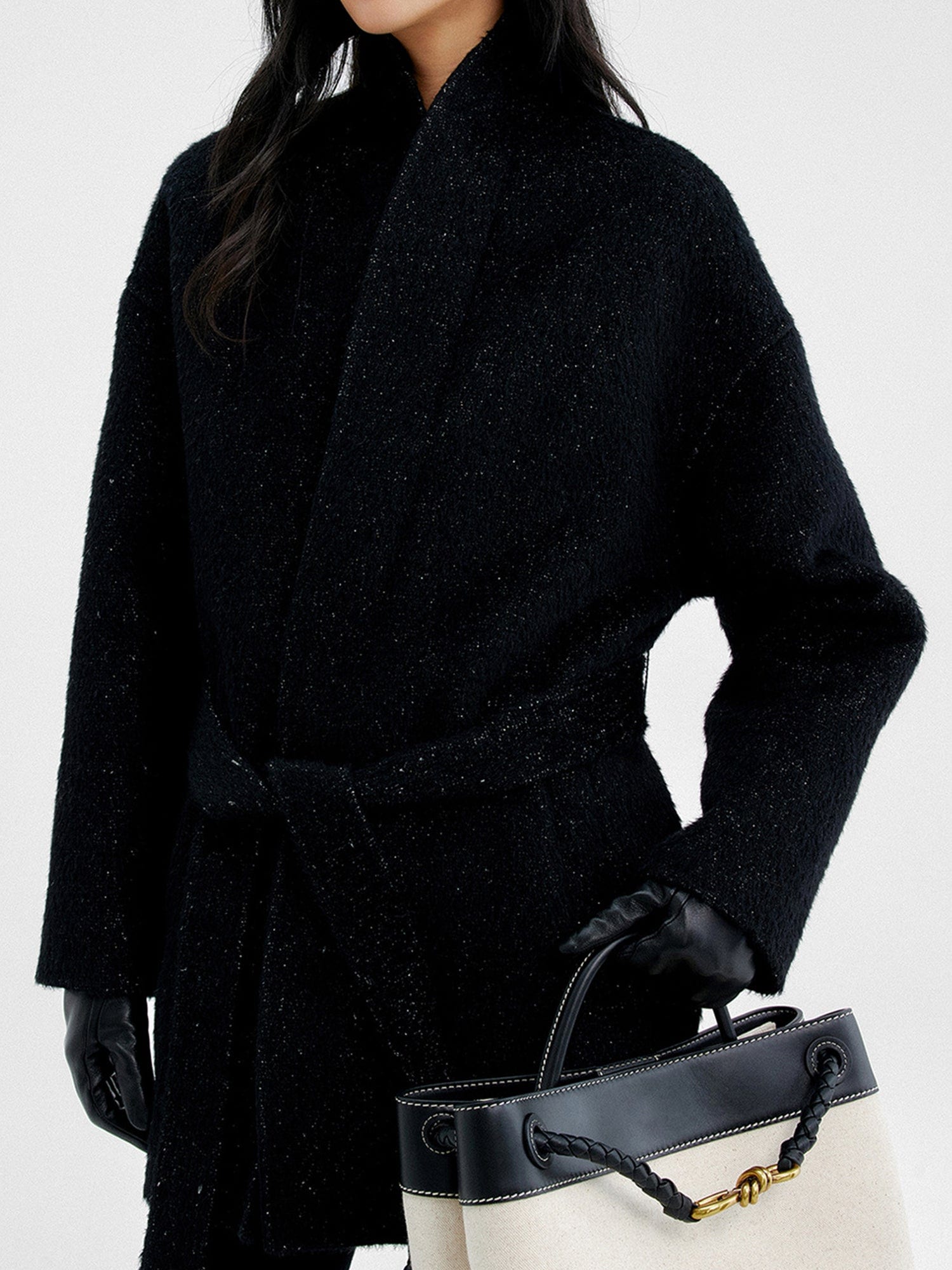 Woman wearing Arkin Studio metallic trim belted wool coat in black and black gloves, holding a white bag, standing sideways, on a white background