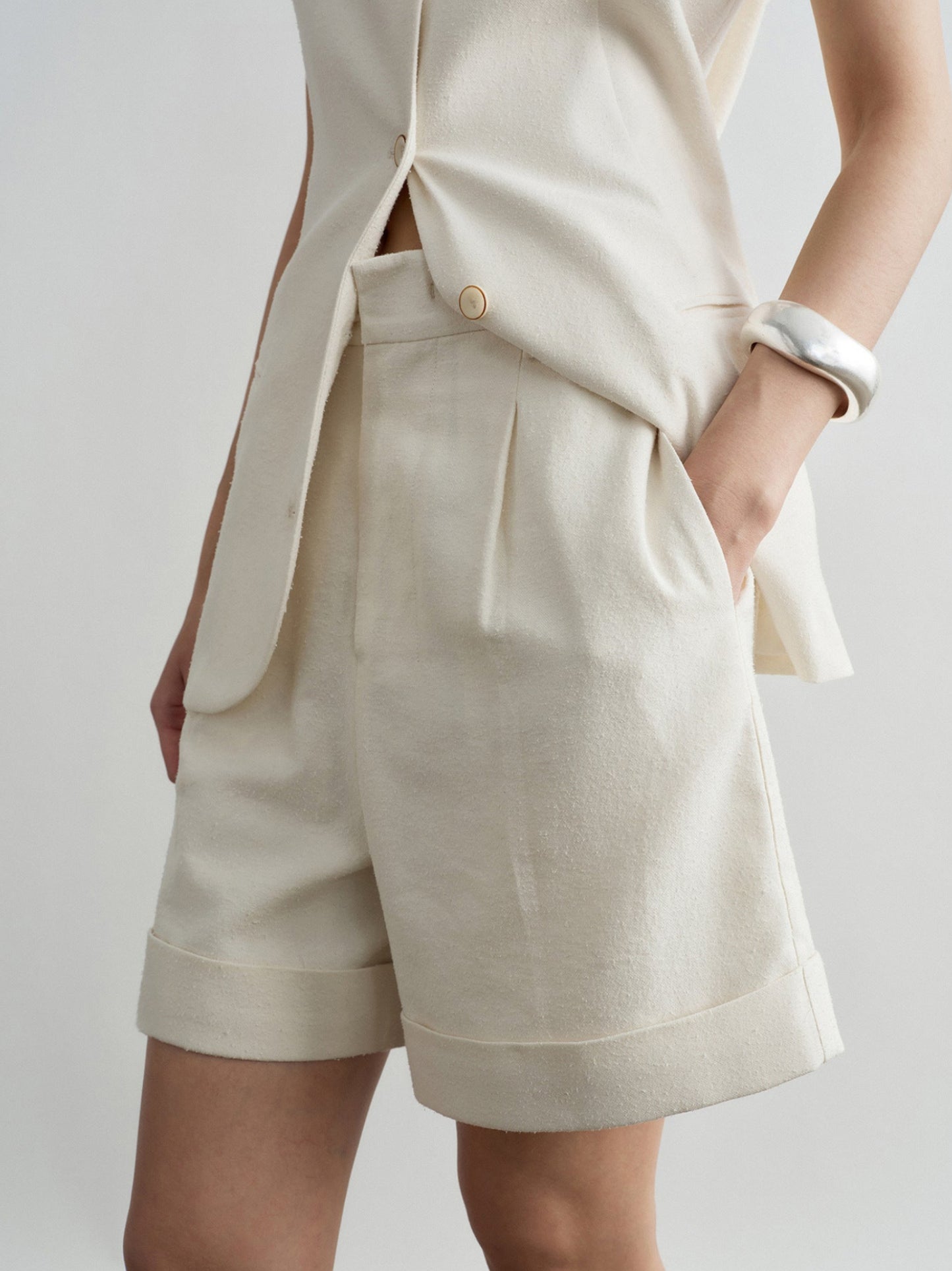 Woman wearing Arkin Studio minimalist cuffed a-line shorts in ivory and matching sleeveless top, one hand in the shorts' pocket, standing sideways, on a white background
