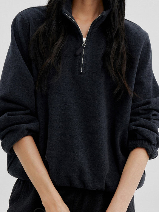 Woman wearing Arkin Studio minimalist half zip sweatshirt in charcoal, with both sleeves half-rolled up on her forearms, on a white background