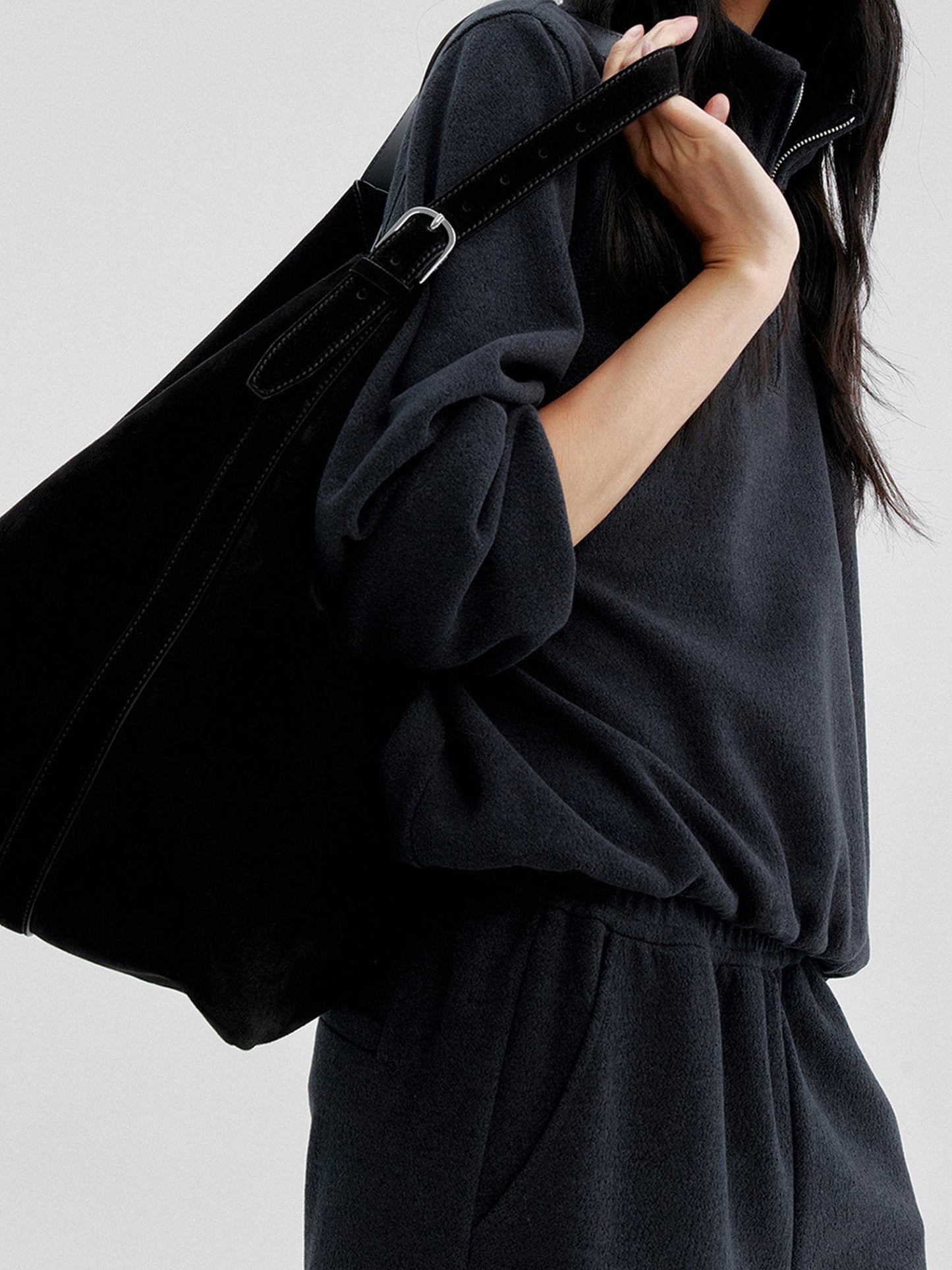 Woman wearing Arkin Studio minimalist half zip sweatshirt in charcoal, with one sleeve half-rolled up on her right forearm, a black bag on her back, standing sideways, on a white background