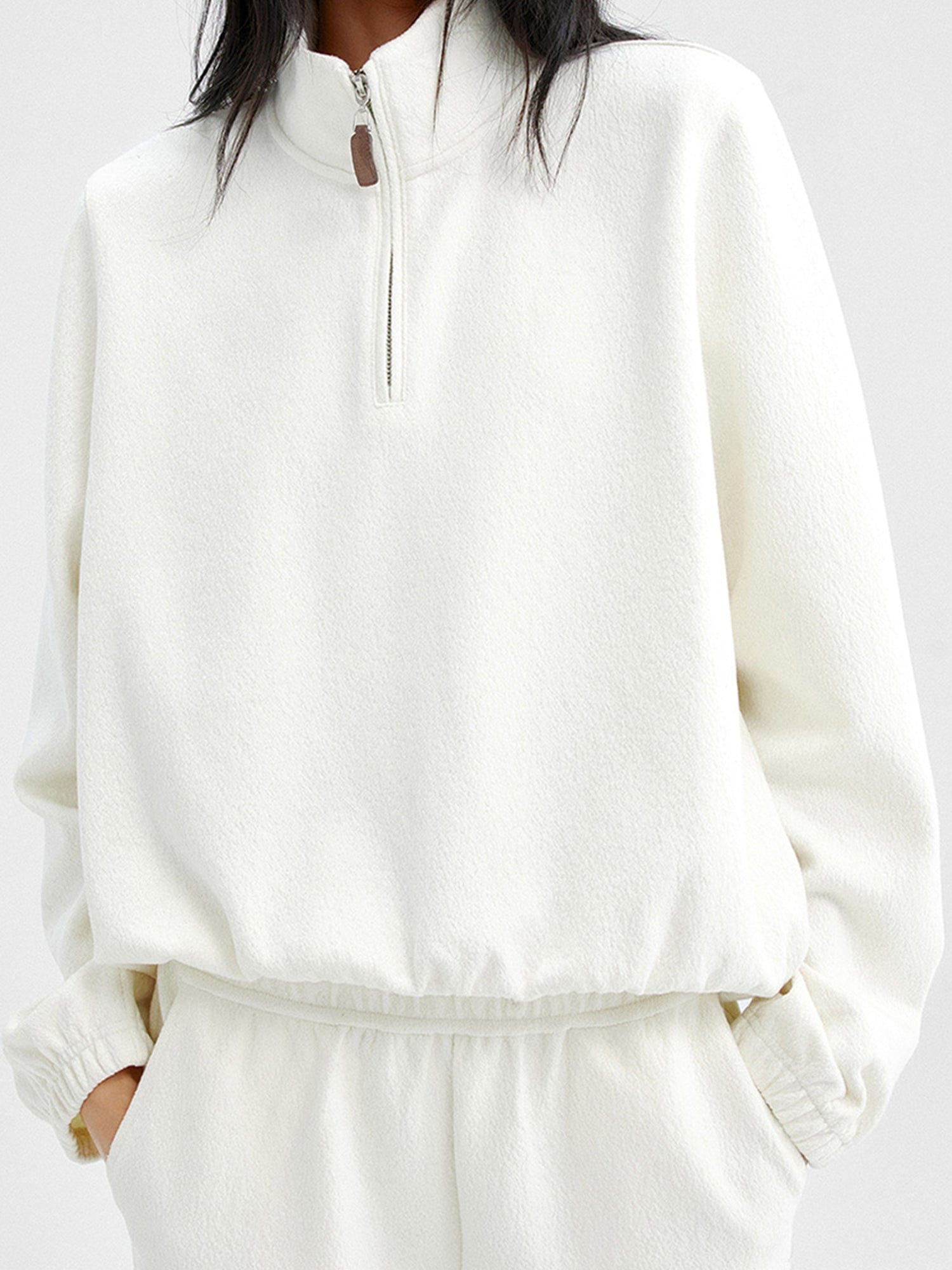 Woman wearing Arkin Studio minimalist half zip sweatshirt in ivory and matching pants, with both hands in pockets, standing facing the camera, on a white background