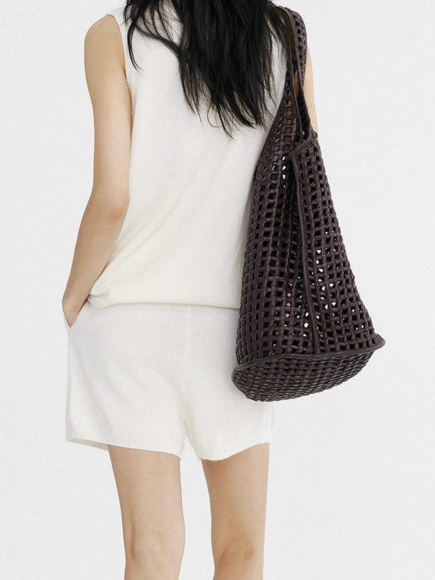 Woman wearing Arkin Studio minimalist round neck tank top in white and matching shorts with a brown bag on her right shoulder, hands in pockets, standing facing a white wall, on a white background