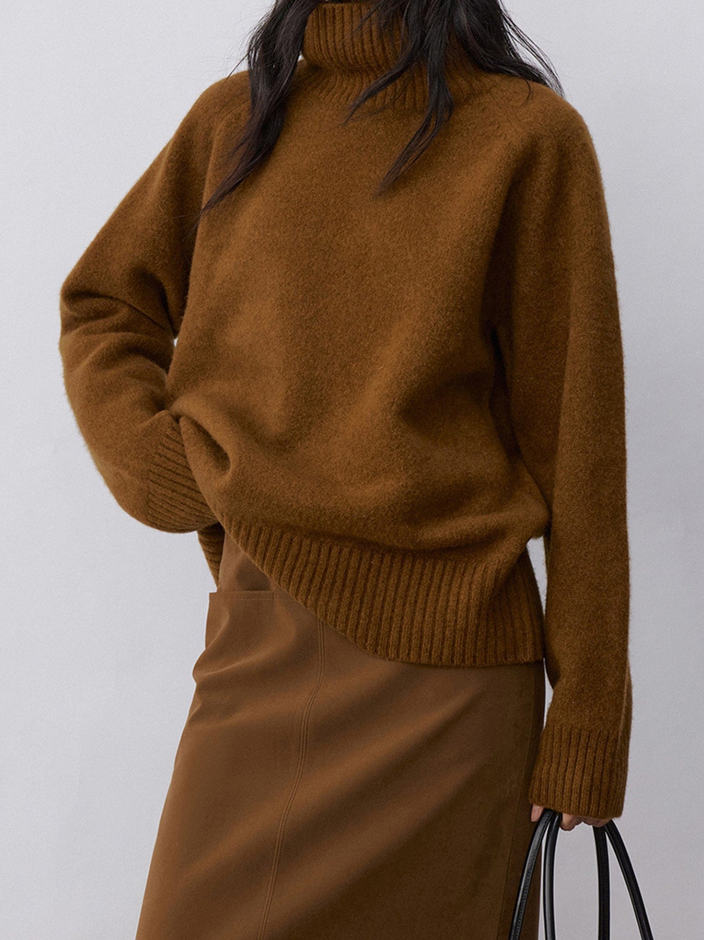 Woman wearing Arkin Studio minimalist turtleneck wool sweater in brown and matching skirt, one hand is holding a bag, another is touching the waist, standing facing the camera, on a white background