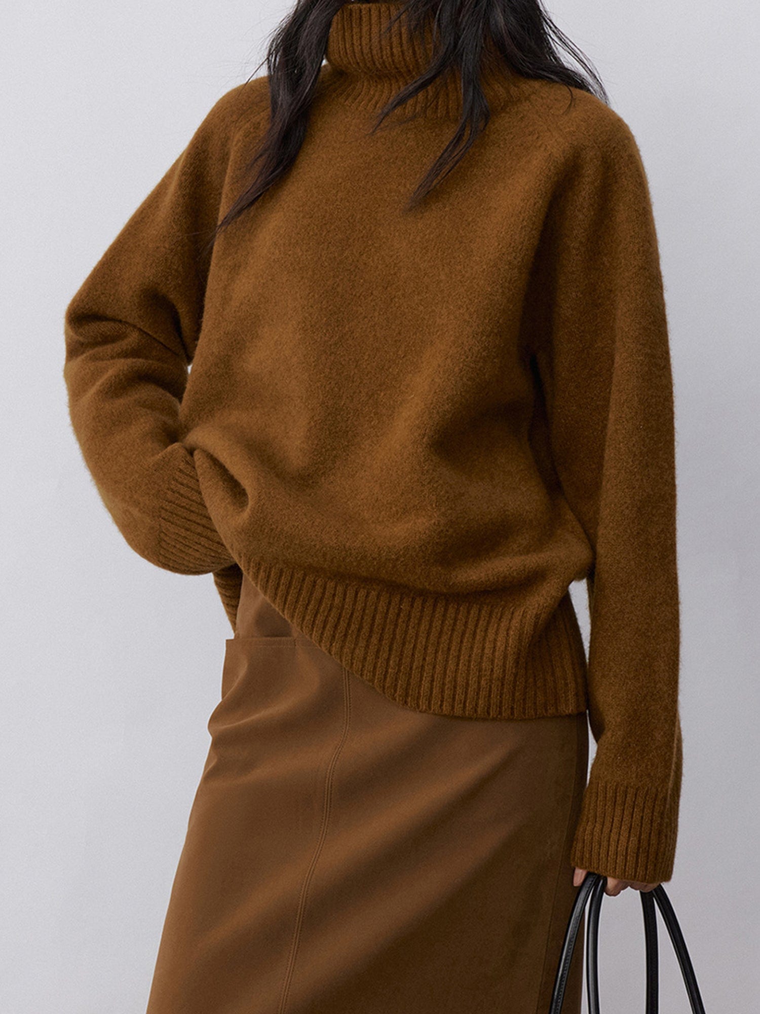Woman wearing Arkin Studio minimalist turtleneck wool sweater in brown and matching skirt, one hand is holding a bag, another is touching the waist, standing facing the camera, on a white background