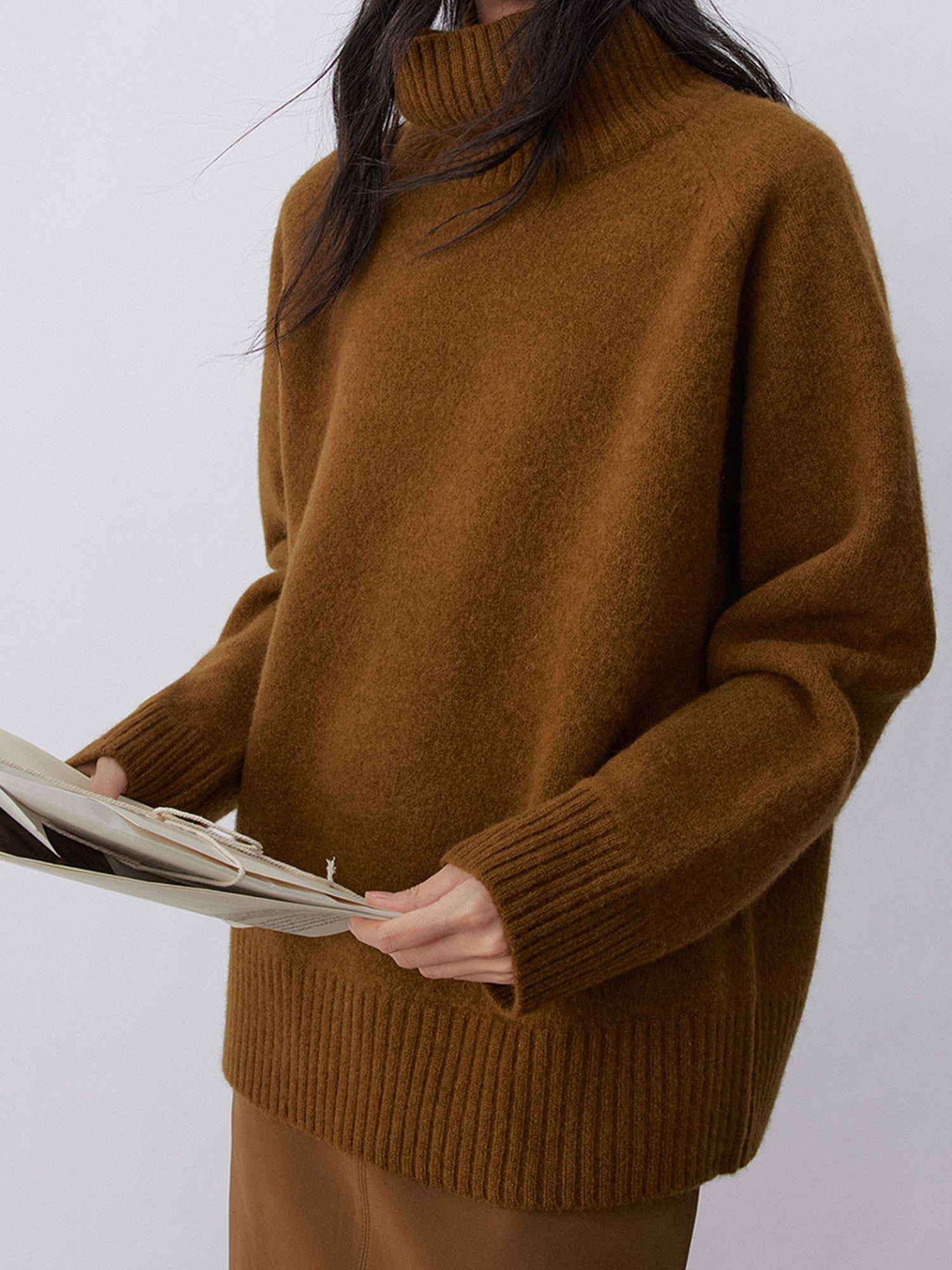 Woman wearing Arkin Studio minimalist turtleneck wool sweater in brown and matching skirt, holding a book, standing sideways, on a white background