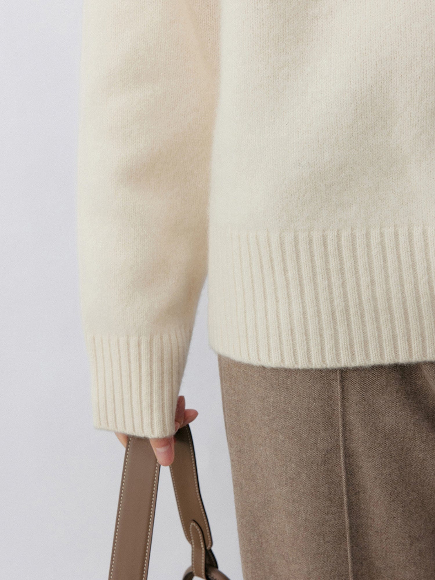 Close up of a woman wearing Arkin Studio minimalist turtleneck wool sweater in white, matching taupe trousers, focusing on her hand holding a white bag, standing facing the camera, on a white background