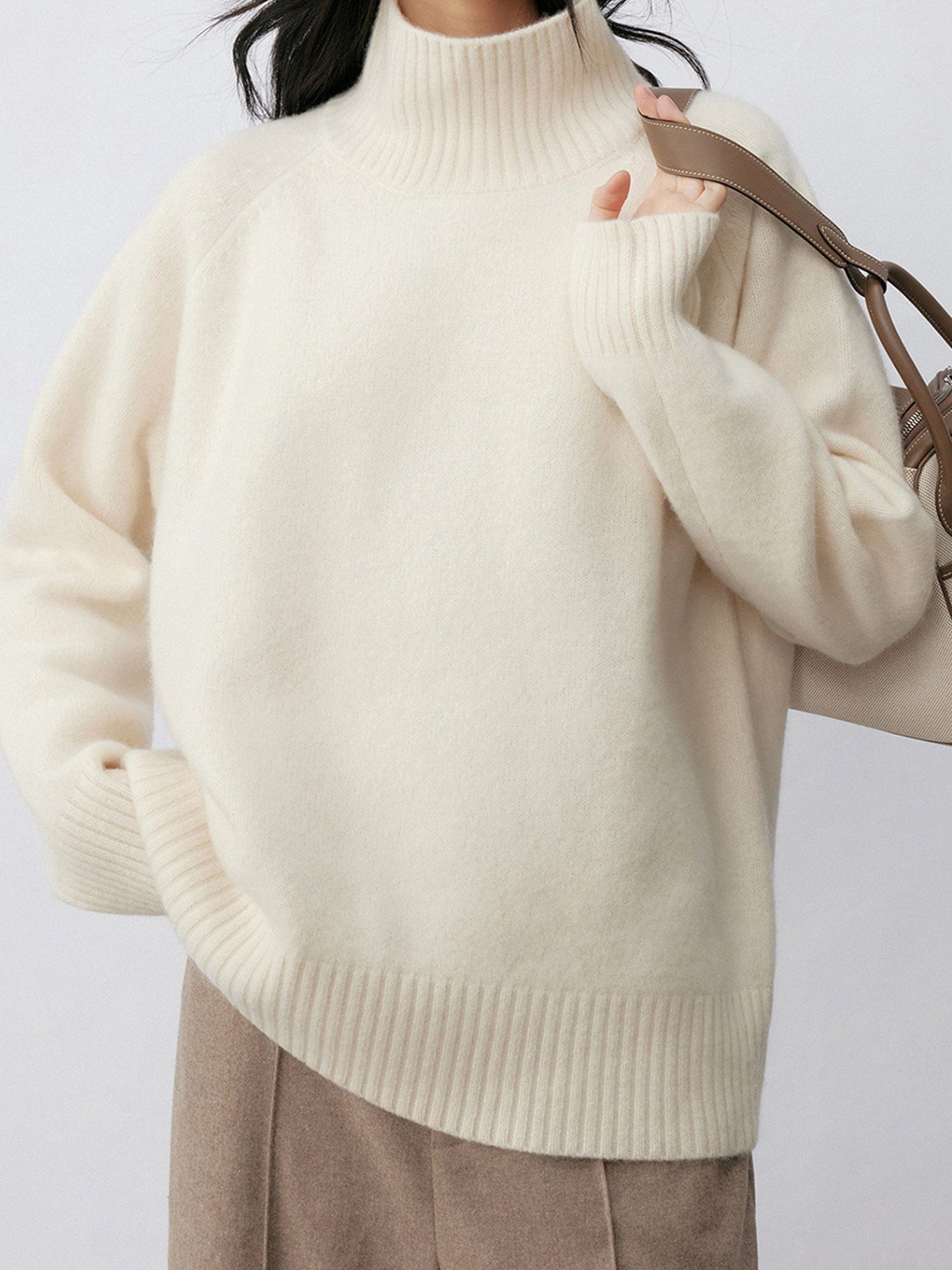 Woman wearing Arkin Studio minimalist turtleneck wool sweater in white and taupe pants, with a white bag on her left shoulder, standing facing the camera, on a white background
