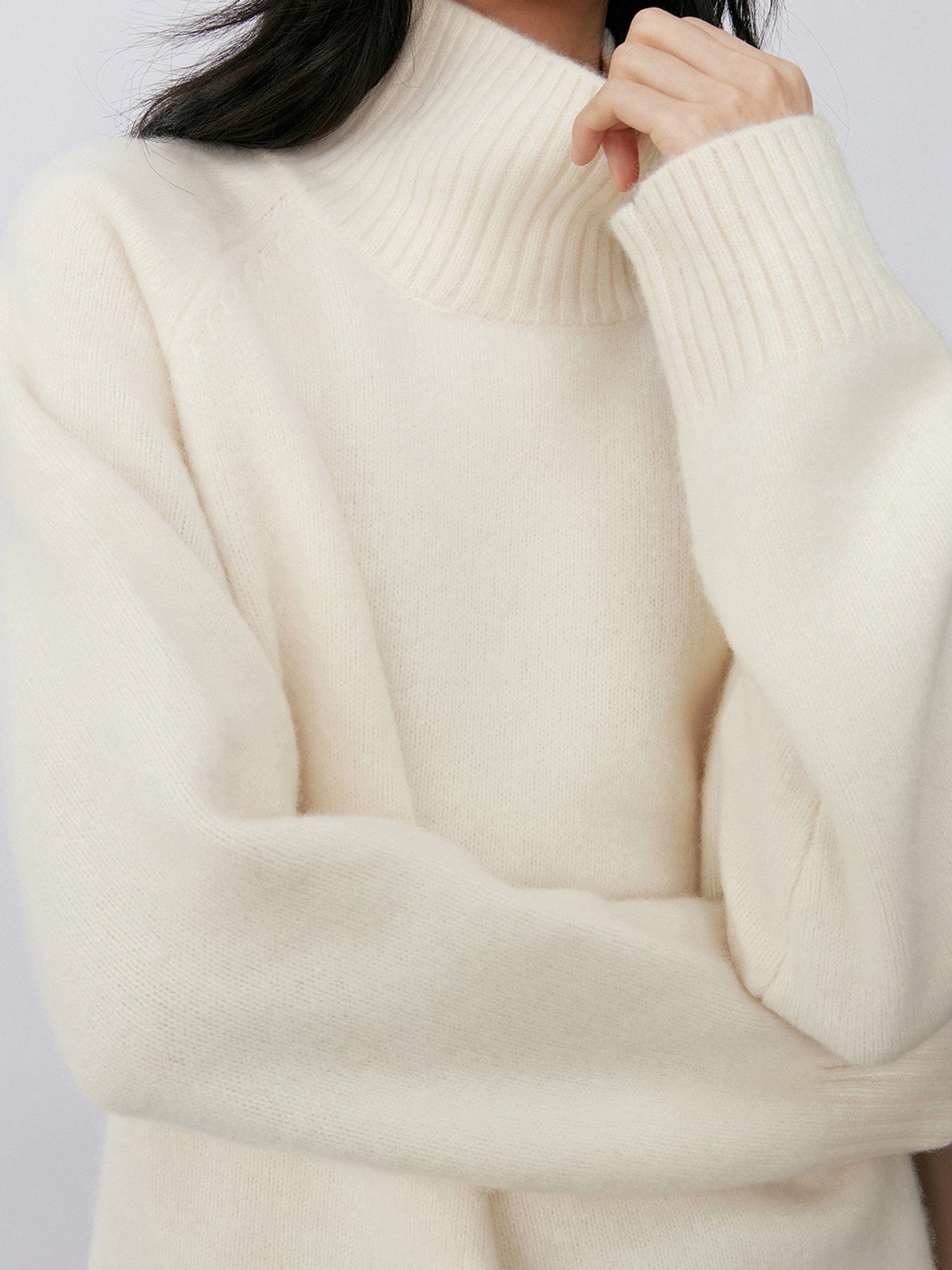 Woman wearing Arkin Studio minimalist turtleneck wool sweater in white, one hand touching the turtleneck, standing sideways, on a white background